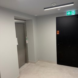 NSB BUILD sp. z o.o. - Wnętrze korytarza z windą i czarnymi drzwiami ewakuacyjnymi, oświetlonego ledowymi lampami. Jasne ściany i podłoga z płytek. Widoczny znak ewakuacyjny.