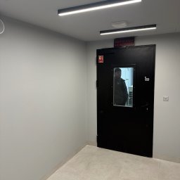 NSB BUILD sp. z o.o. - Wnętrze biurowe z czarnymi drzwiami z okienkiem, przez które widać osobę. Nad drzwiami tabliczka informacyjna i oświetlenie LED. Jasne ściany i podłoga z płytek.