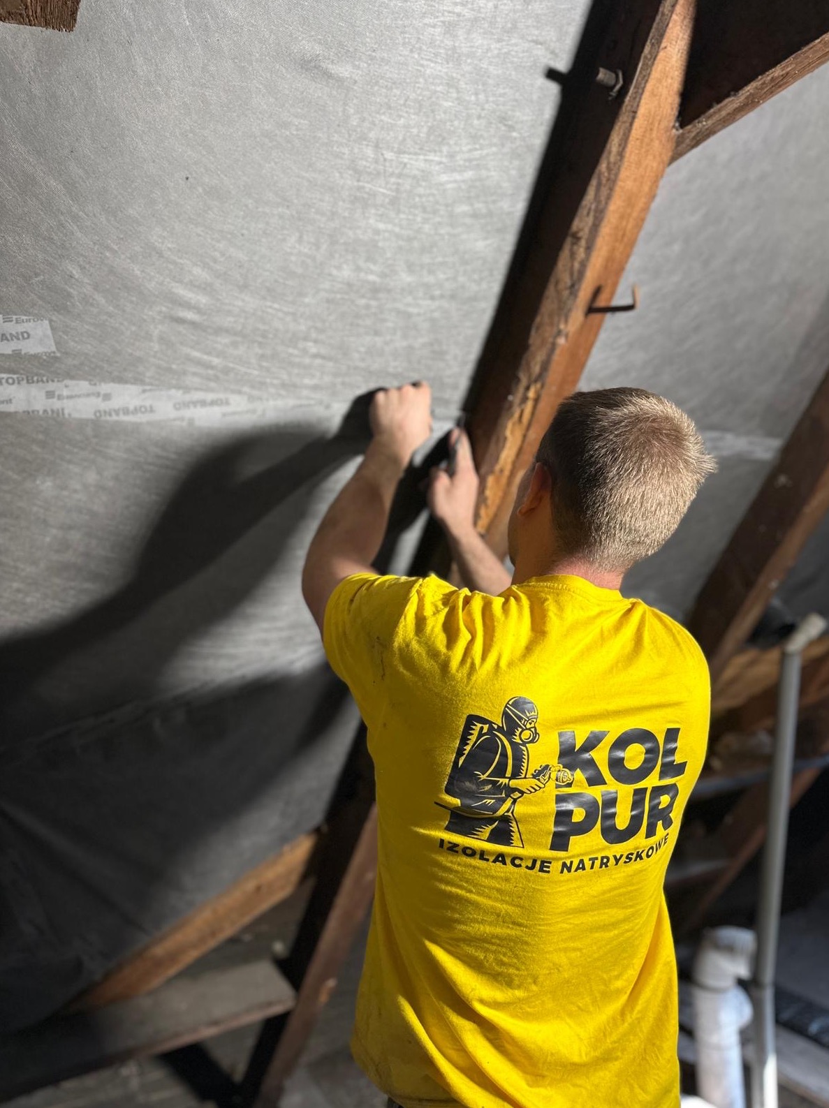 Pracownik w żółtej koszulce z logo firmy montuje szarą membranę izolacyjną na drewnianej konstrukcji dachu poddasza. Widoczny etap przygotowania pod ocieplenie.