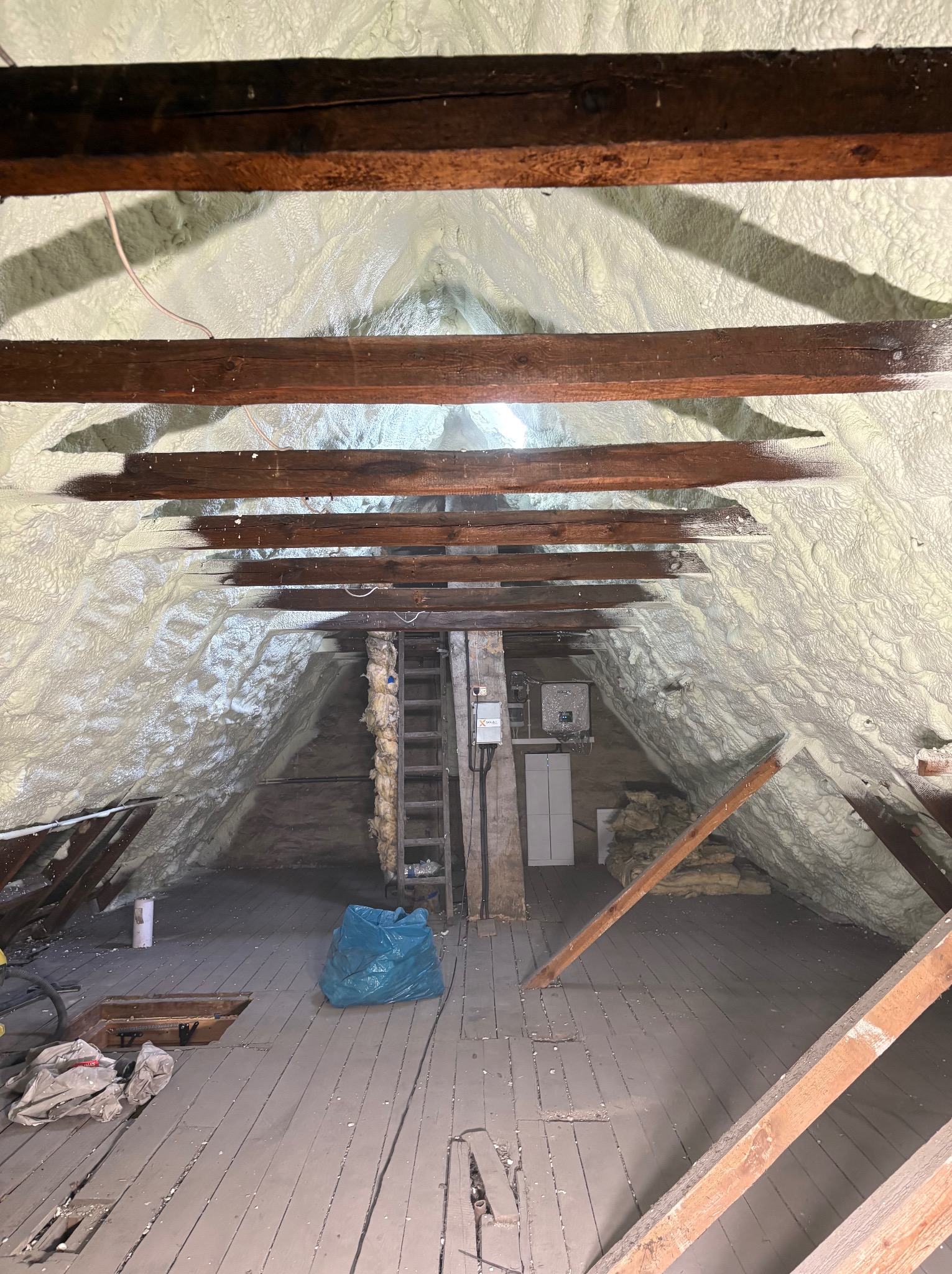 Poddasze z drewnianą konstrukcją, pokryte pianką izolacyjną, z widocznymi elementami instalacji i workiem na odpady, ukazujące etap prac ociepleniowych.