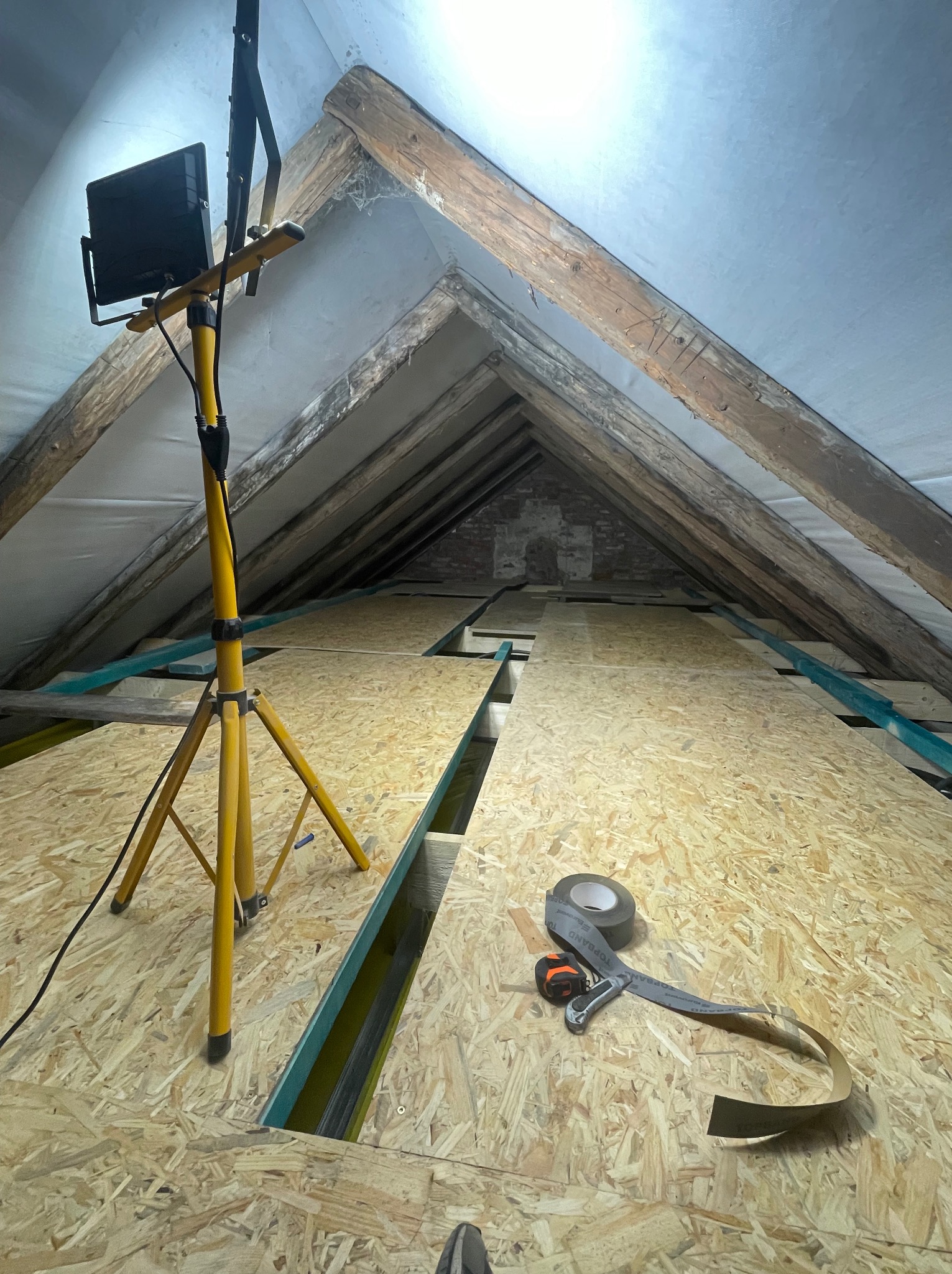 Poddasze z drewnianą konstrukcją dachu, oświetlone lampą na statywie. Częściowo ułożona podłoga z płyt OSB, taśma uszczelniająca i miarka leżą na podłodze.