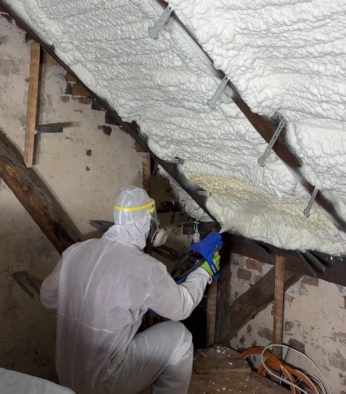 Pracownik w kombinezonie ochronnym aplikuje piankę PUR na poddasze z widocznymi elementami więźby dachowej i fragmentem ściany z cegły.