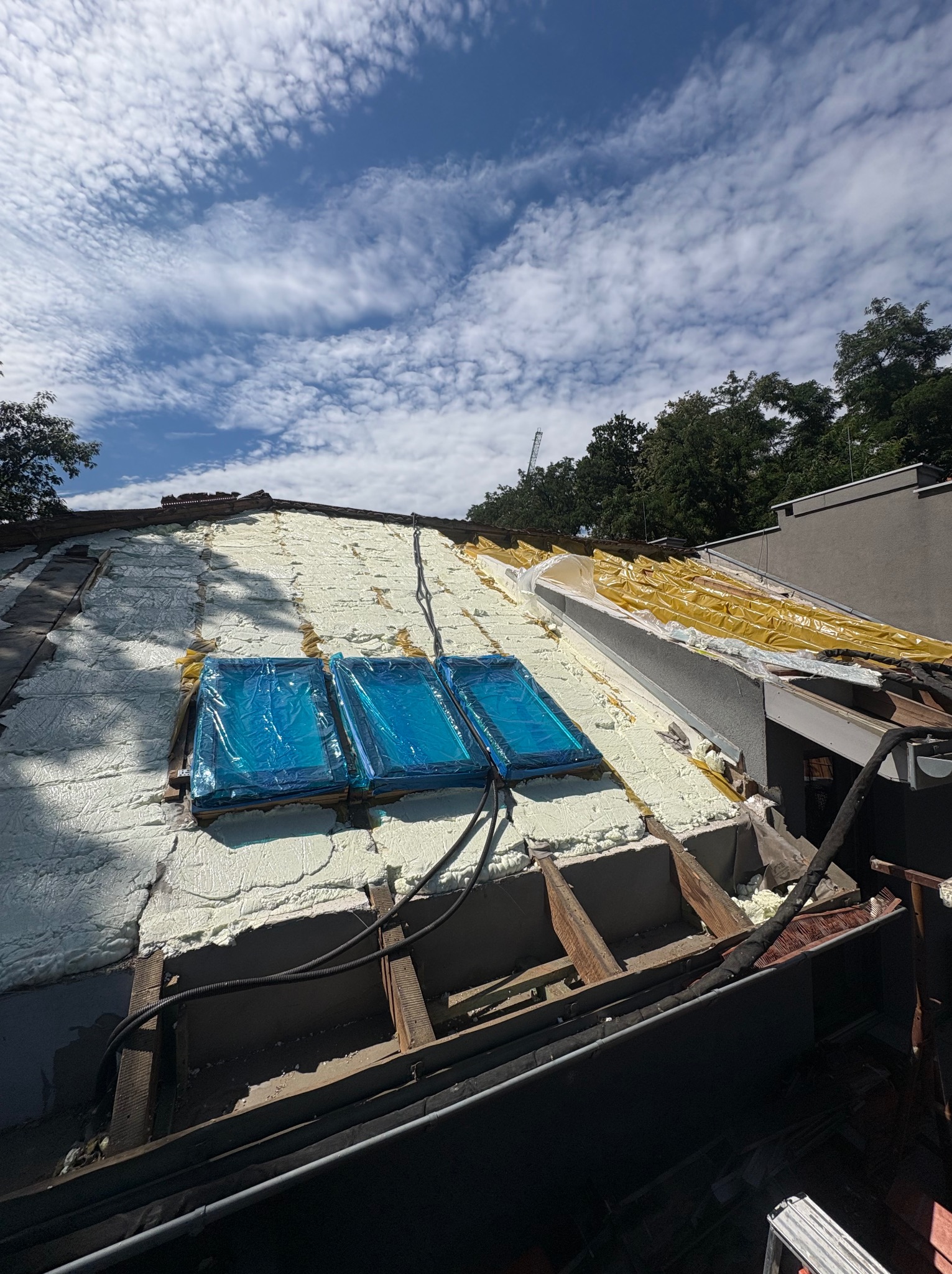 Ocieplenie poddasza pianką PUR w trakcie realizacji, widoczne fragmenty drewnianej konstrukcji dachu i zabezpieczone okna dachowe. Błękitne niebo w tle.