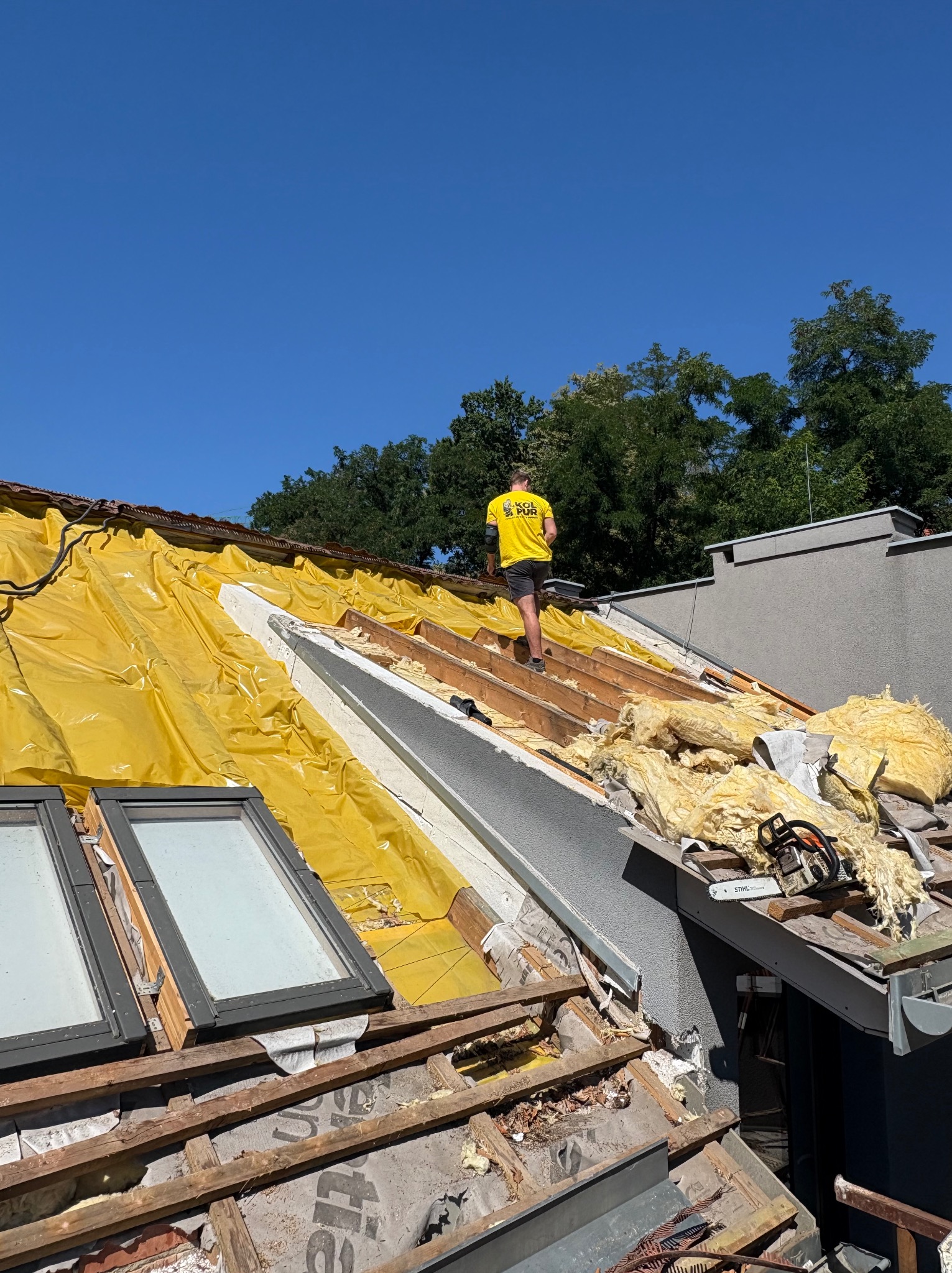 Prace na dachu: żółta folia, wełna mineralna, okna dachowe. Pracownik w firmowej koszulce porusza się po konstrukcji. Widoczne narzędzia i elementy izolacji.