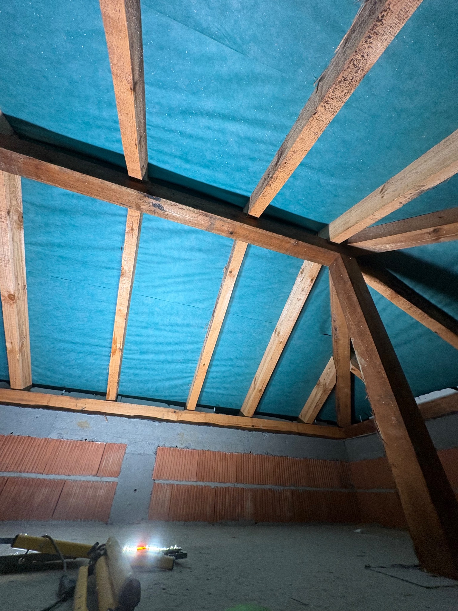 Poddasze z drewnianą konstrukcją dachu zabezpieczoną niebieską membraną, widoczne ściany z pustaków. Przy podłodze narzędzia w słabym oświetleniu.