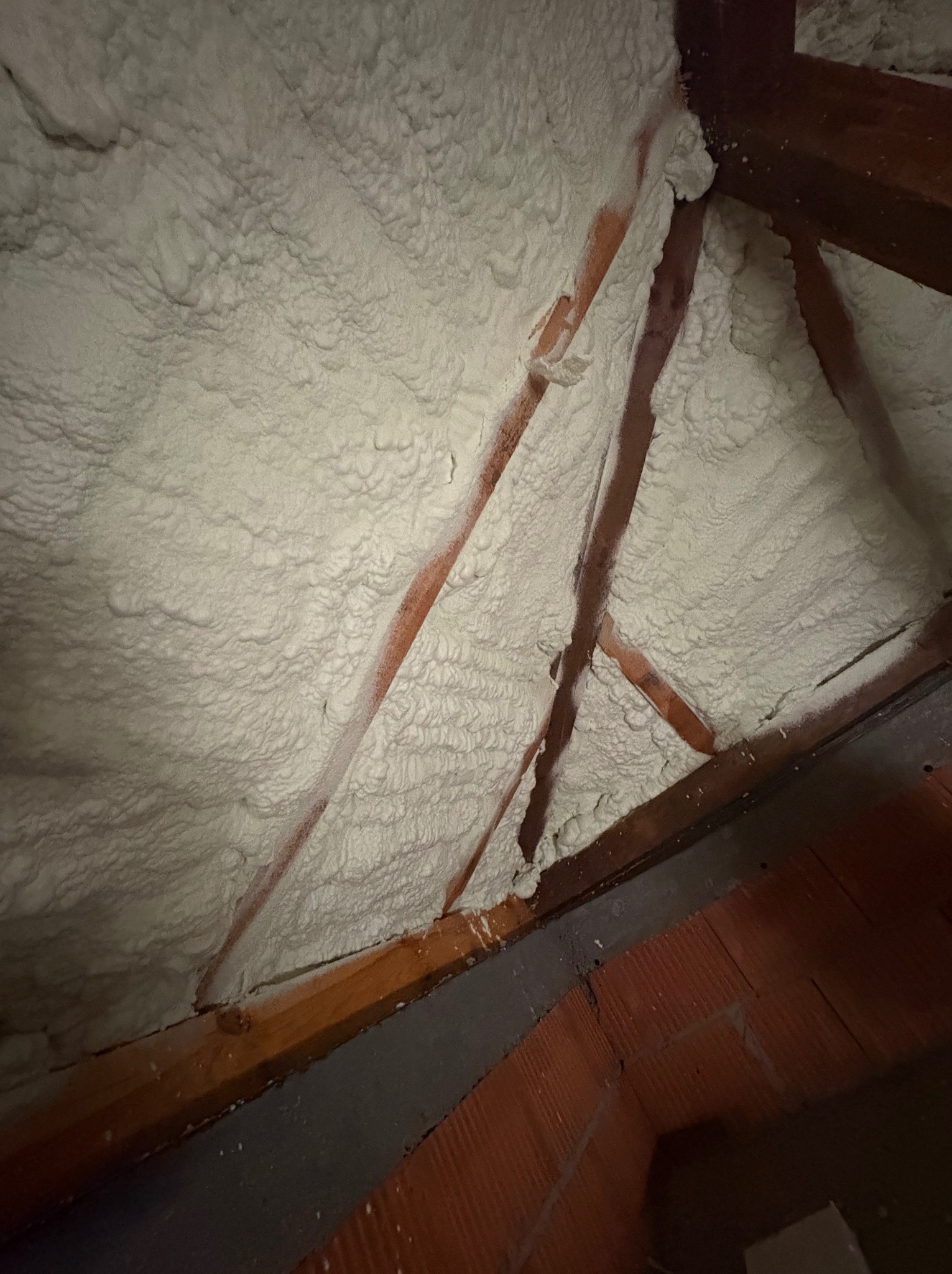 Poddasze z drewnianą konstrukcją, ocieplone grubą warstwą pianki PUR o nieregularnej fakturze, widoczne fragmenty ceglanej ściany w dolnej części kadru.