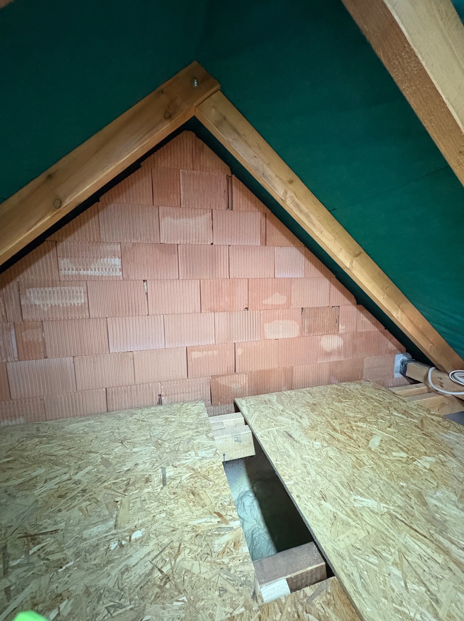 Poddasze z ceglaną ścianą szczytową, deskami podłogowymi OSB i zieloną membraną dachową. Widoczna izolacja w przestrzeni pod deskami.