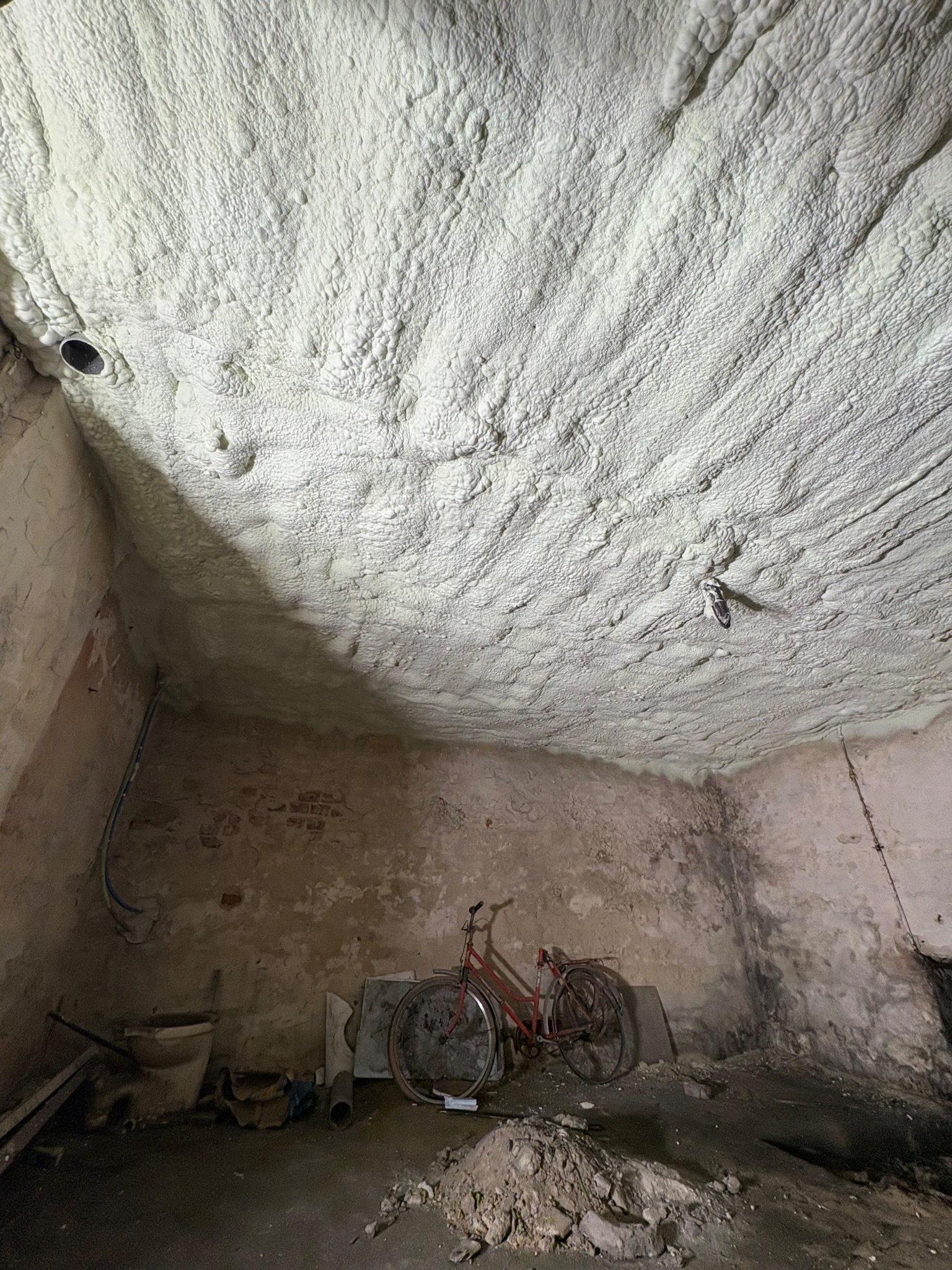 Pomieszczenie z grubą warstwą białej pianki izolacyjnej na suficie, rower oparty o ścianę, widoczne ślady wilgoci i zabrudzeń. Nietypowe ujęcie z dołu.