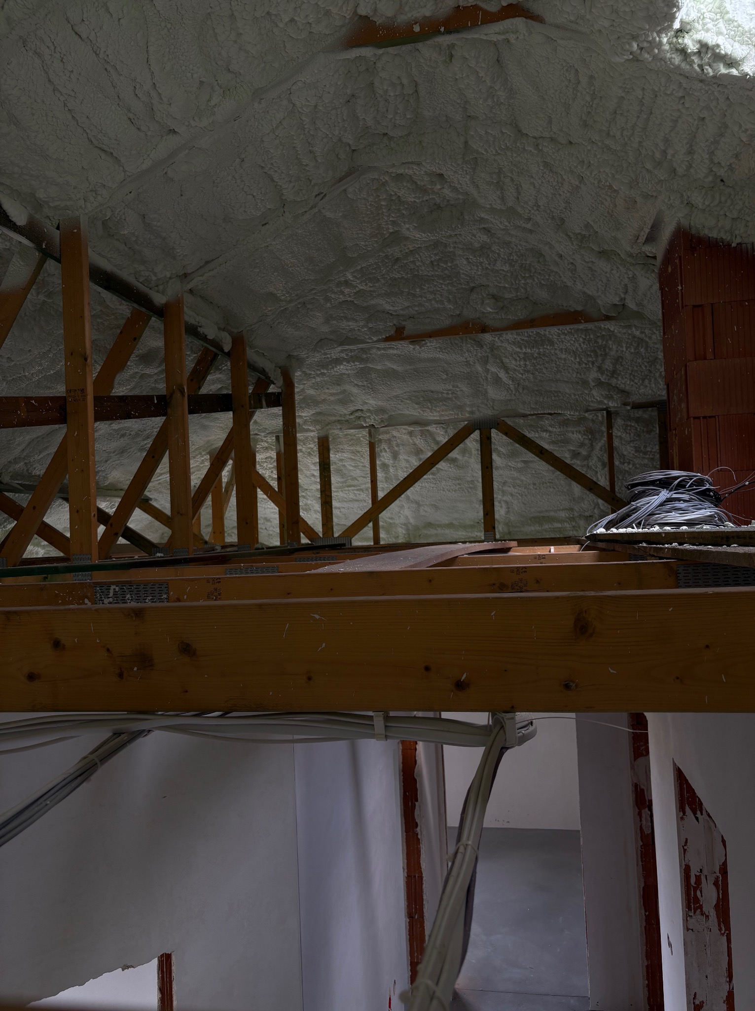 Poddasze z drewnianą konstrukcją i izolacją z pianki PUR, widoczne kable i fragment ściany z cegły. Ujęcie z dołu, ukazujące detale izolacji.