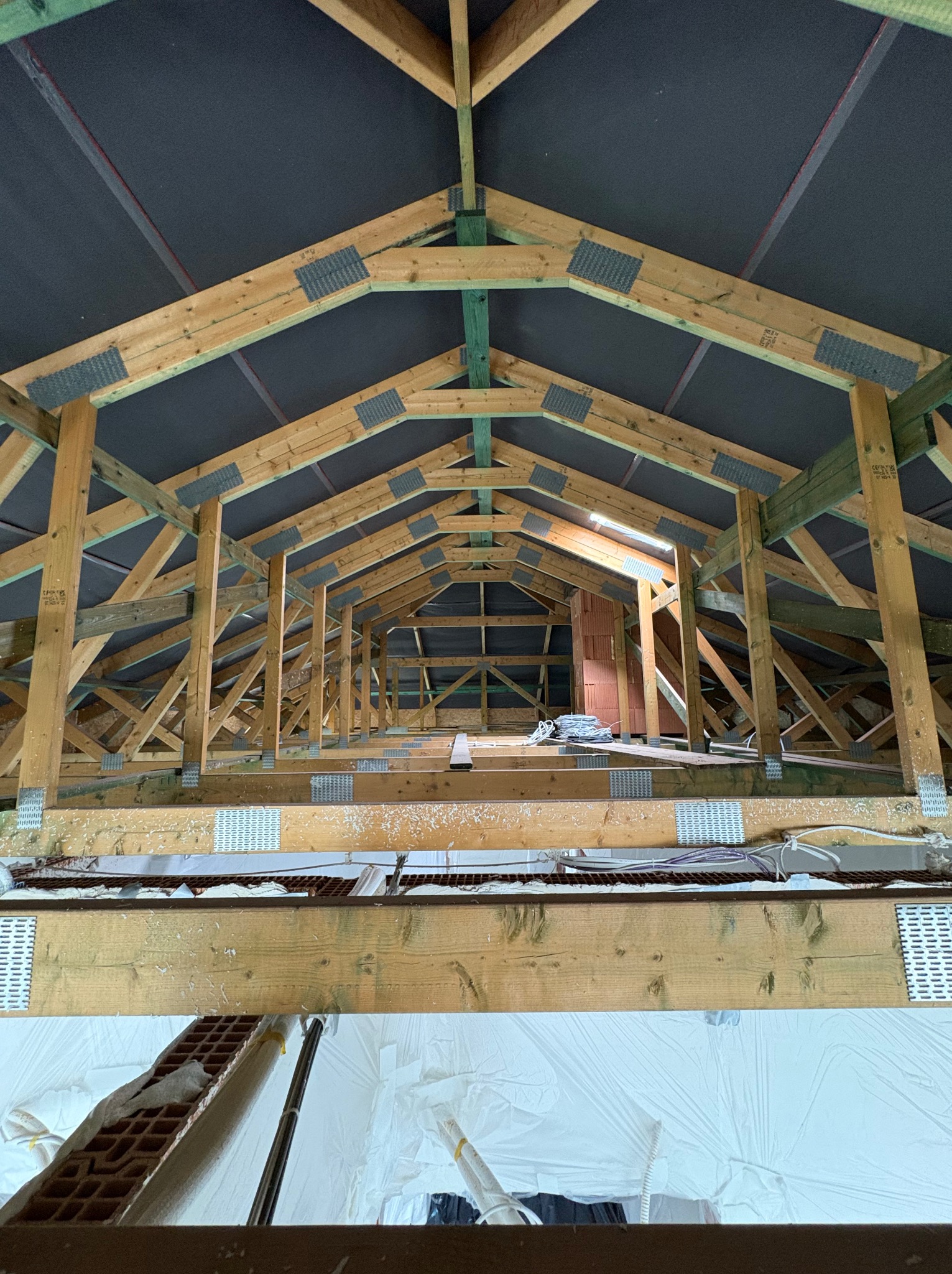 Wnętrze drewnianej konstrukcji dachu z widoczną membraną i elementami izolacji, w trakcie prac. Perspektywa od dołu ukazuje skomplikowaną strukturę więźby dachowej.