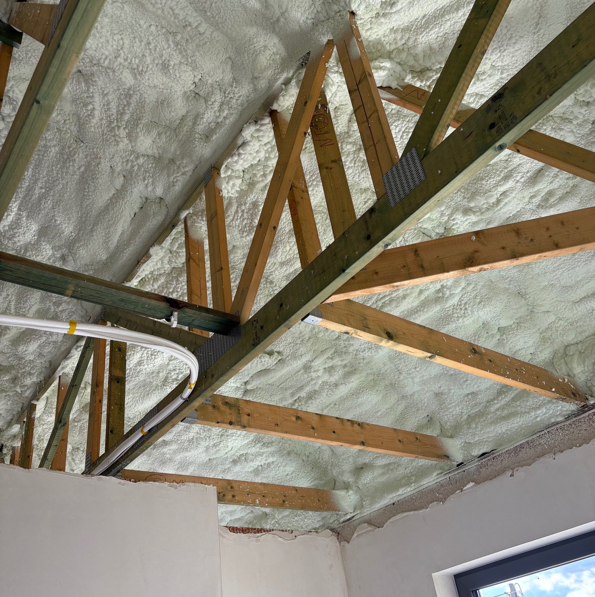 Poddasze z drewnianą konstrukcją dachu i ociepleniem pianką PUR, widoczne przewody elektryczne w osłonie. Detal wykończonej izolacji i surowej ściany.
