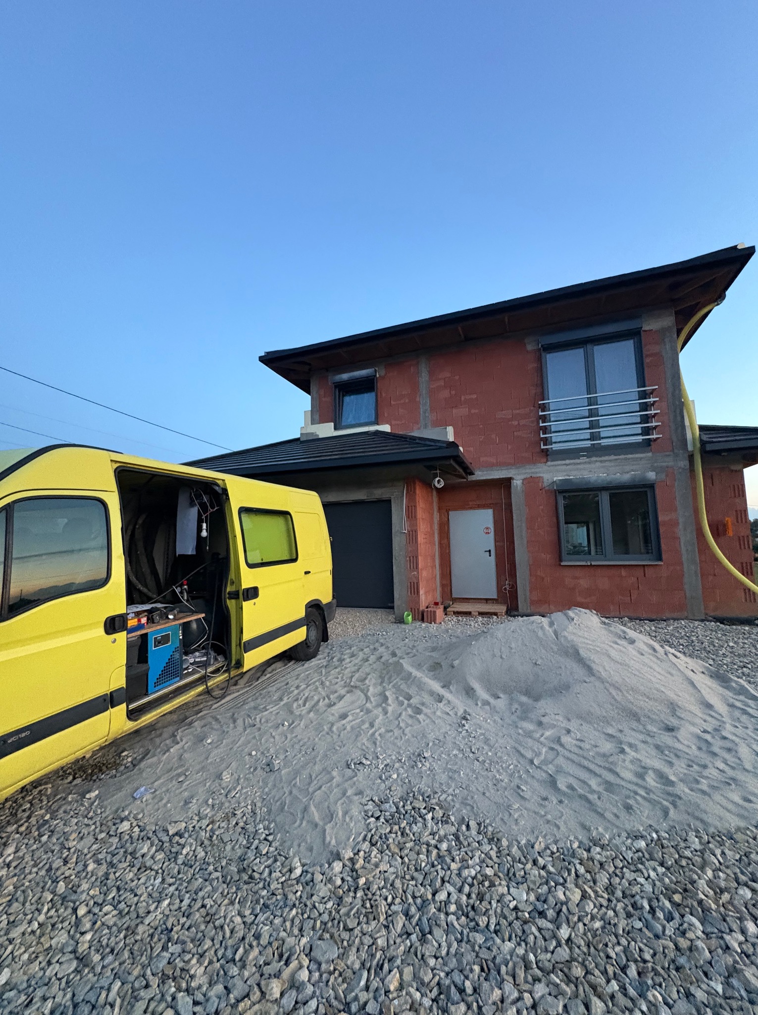 Żółty samochód z otwartymi drzwiami i sprzętem do ocieplania pianką PUR na tle domu w trakcie budowy, z widocznym ociepleniem ścian. Węże i urządzenia.