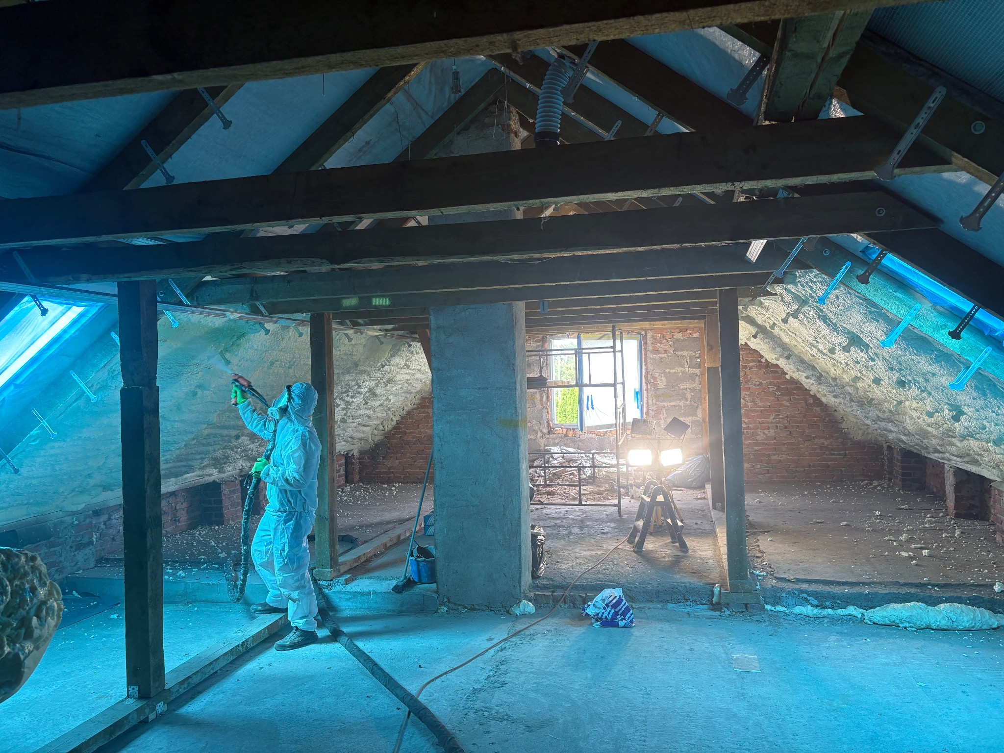 Pracownik w kombinezonie ochronnym natryskuje pianę PUR na poddaszu domu z odsłoniętą cegłą, widoczne drewniane elementy konstrukcyjne i oświetlenie robocze.