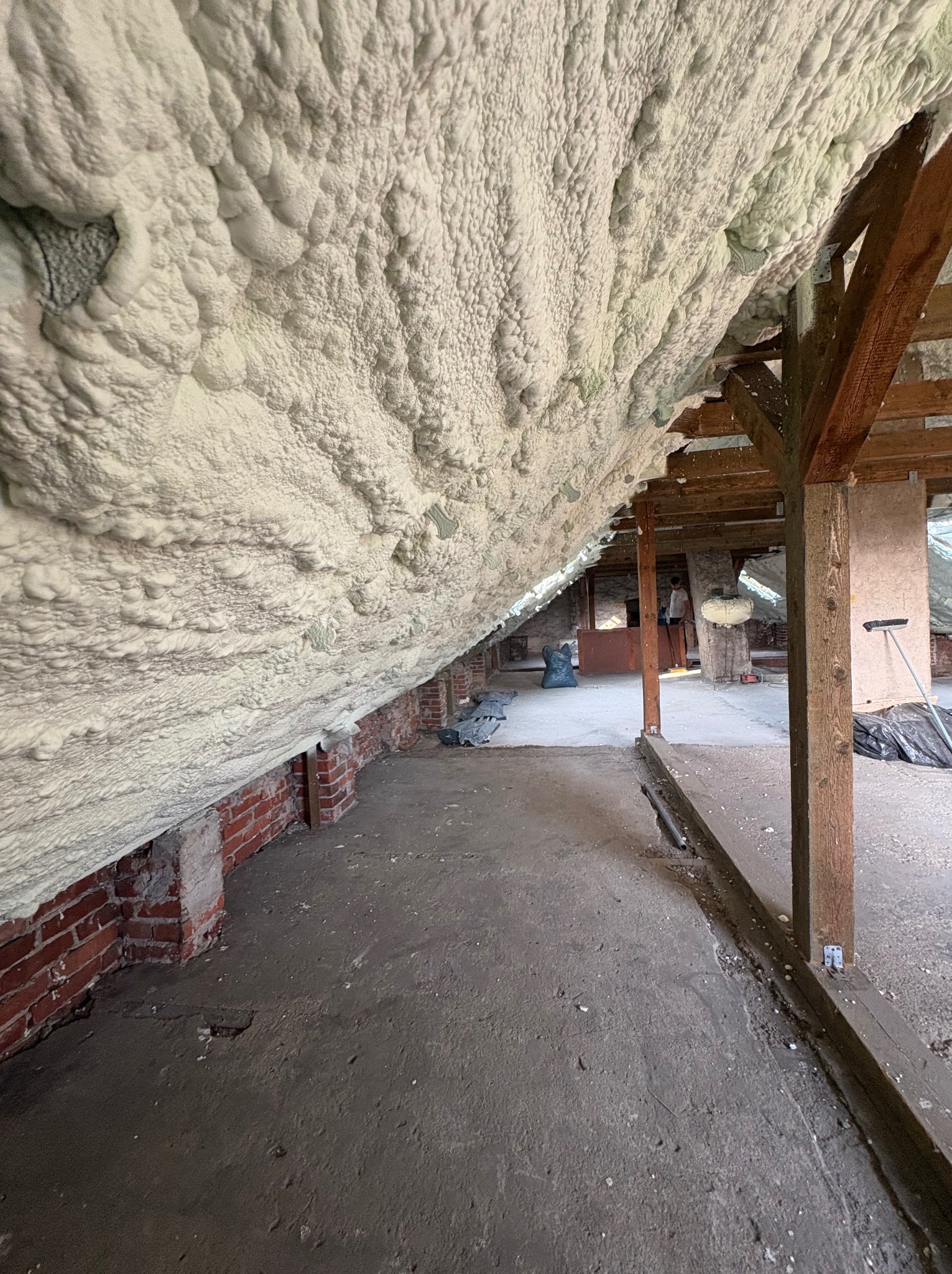 Poddasze po ociepleniu pianką PUR, widoczne krokwie i ceglana ściana. Szara podłoga betonowa. Widok z dołu na izolację poddasza.