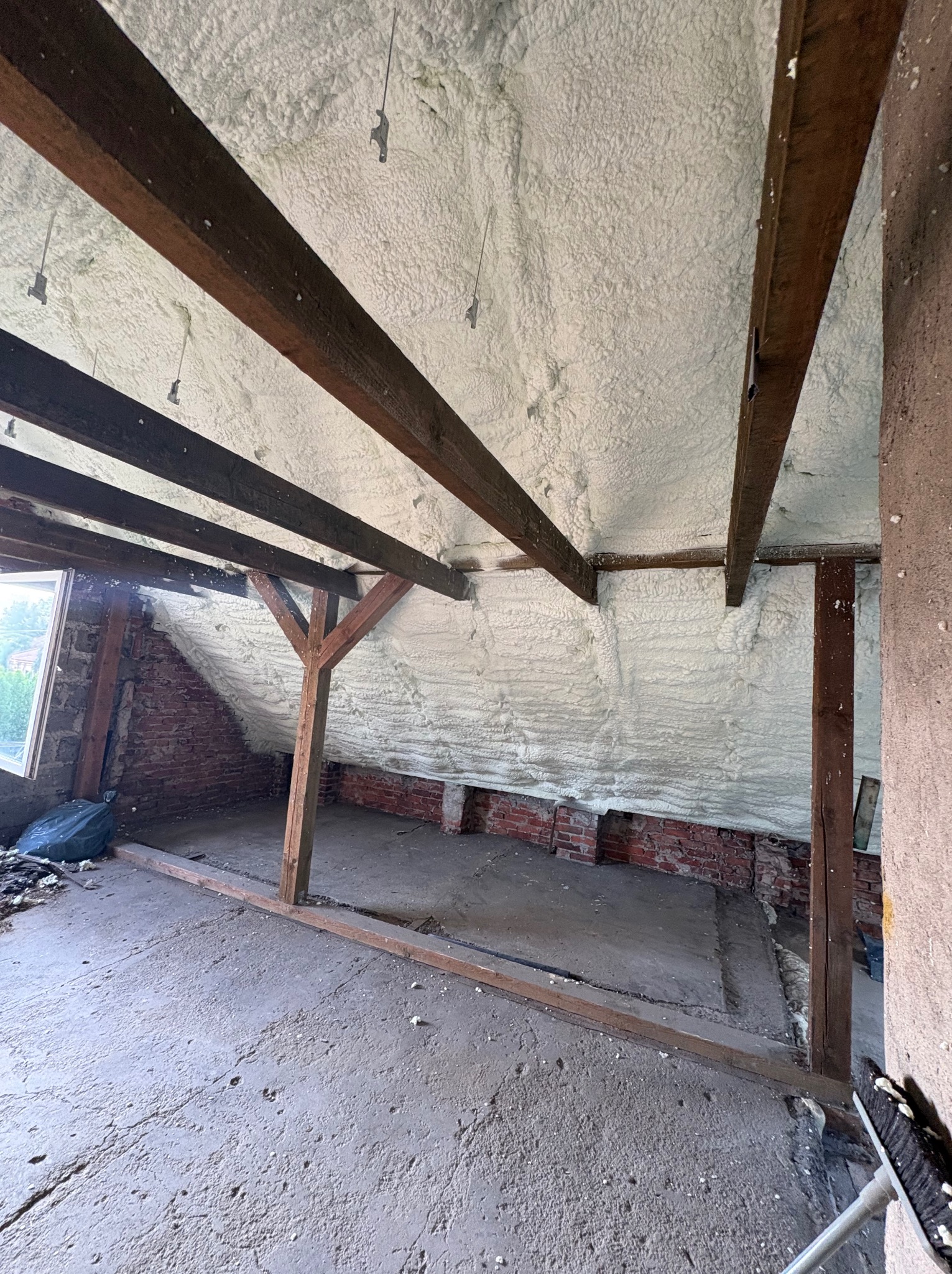 Poddasze po ociepleniu pianką PUR, widoczne drewniane belki i ceglane ściany. Biała izolacja szczelnie wypełnia przestrzeń, tworząc jednolitą warstwę.