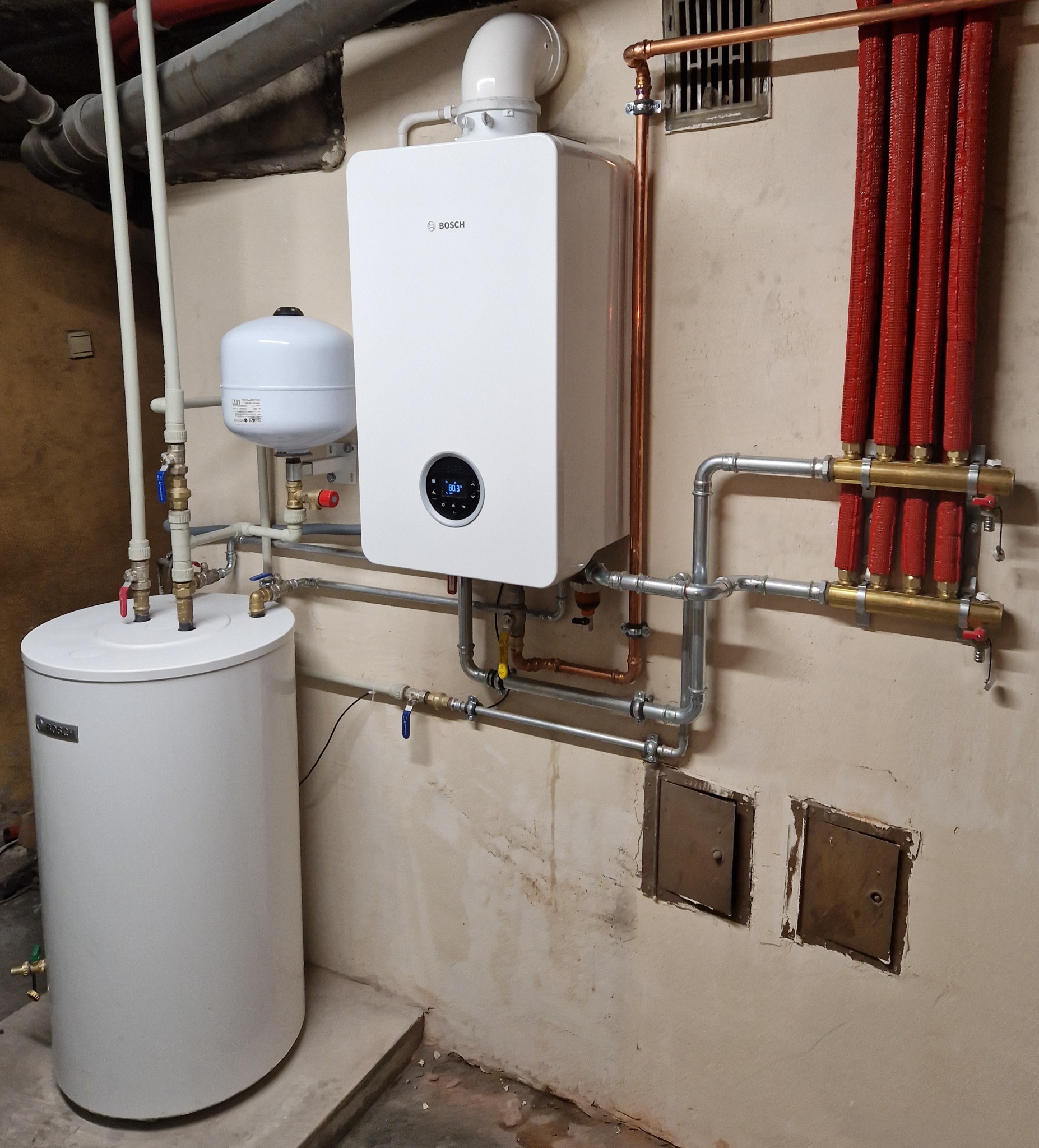 Nowoczesny, biały piec gazowy Bosch z podłączonym zasobnikiem ciepłej wody i rozdzielaczem centralnego ogrzewania z czerwonymi rurami, zamontowany w piwnicy z widocznymi rurami i instalacjami.