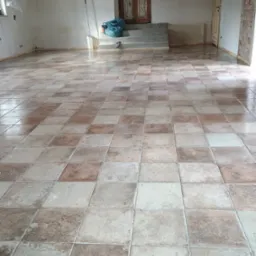 Ułożona podłoga z kwadratowych płytek w odcieniach beżu i brązu, widoczne ślady po remoncie, w tle okno zasłonięte folią i drzwi.