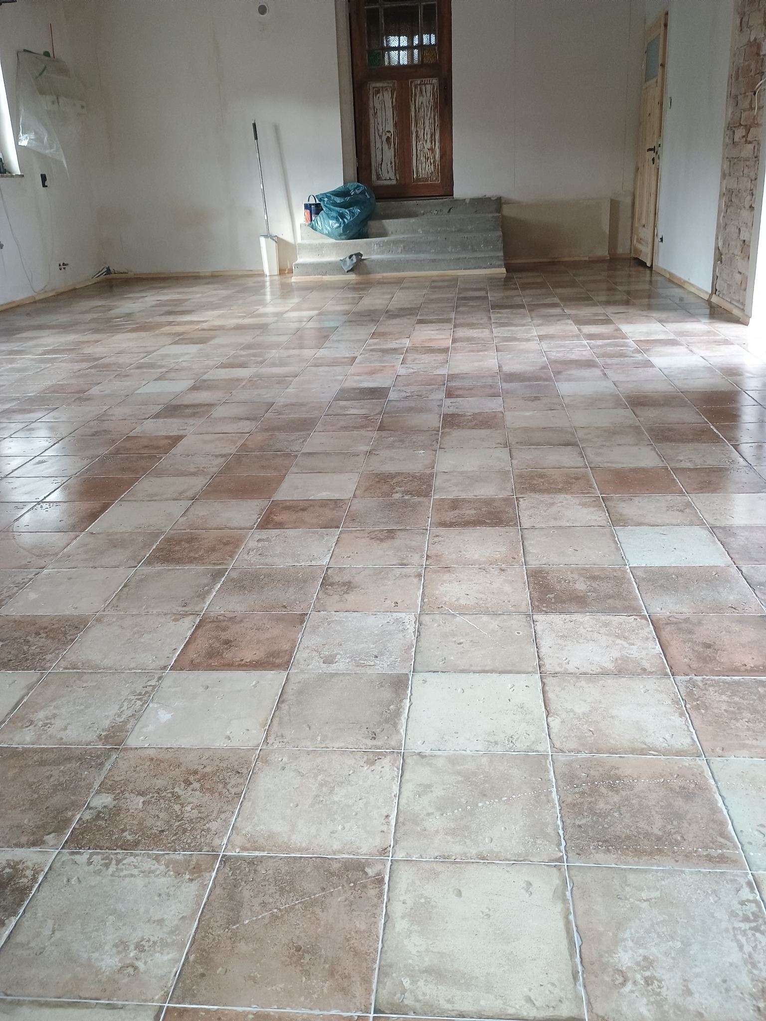 Ułożona podłoga z kwadratowych płytek w odcieniach beżu i brązu, widoczne ślady po remoncie, w tle okno zasłonięte folią i drzwi.