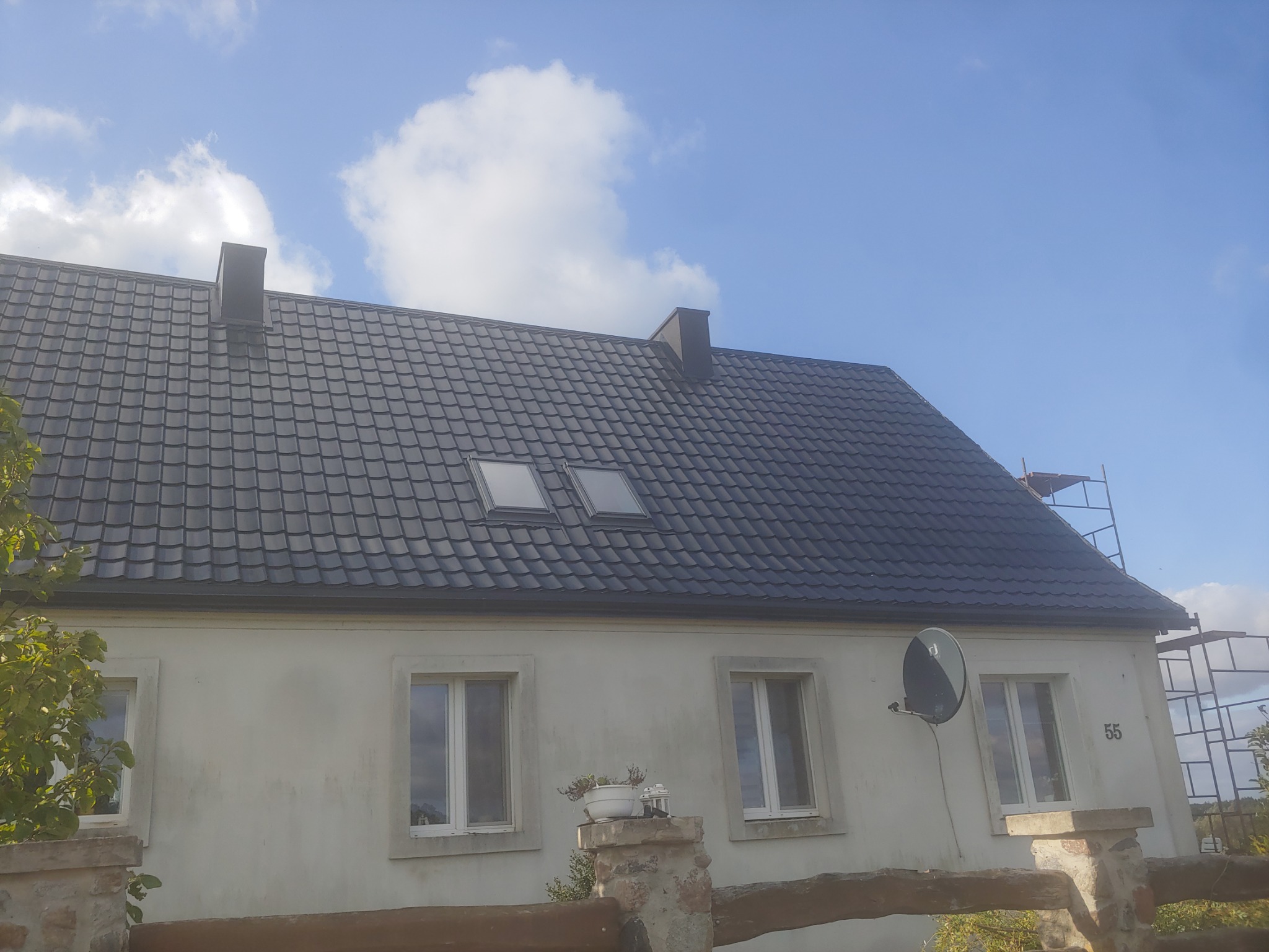 Dom jednorodzinny z nowym, ciemnoszarym dachem z dachówki ceramicznej, widoczne dwa okna dachowe, kominy i rusztowanie po prawej stronie.