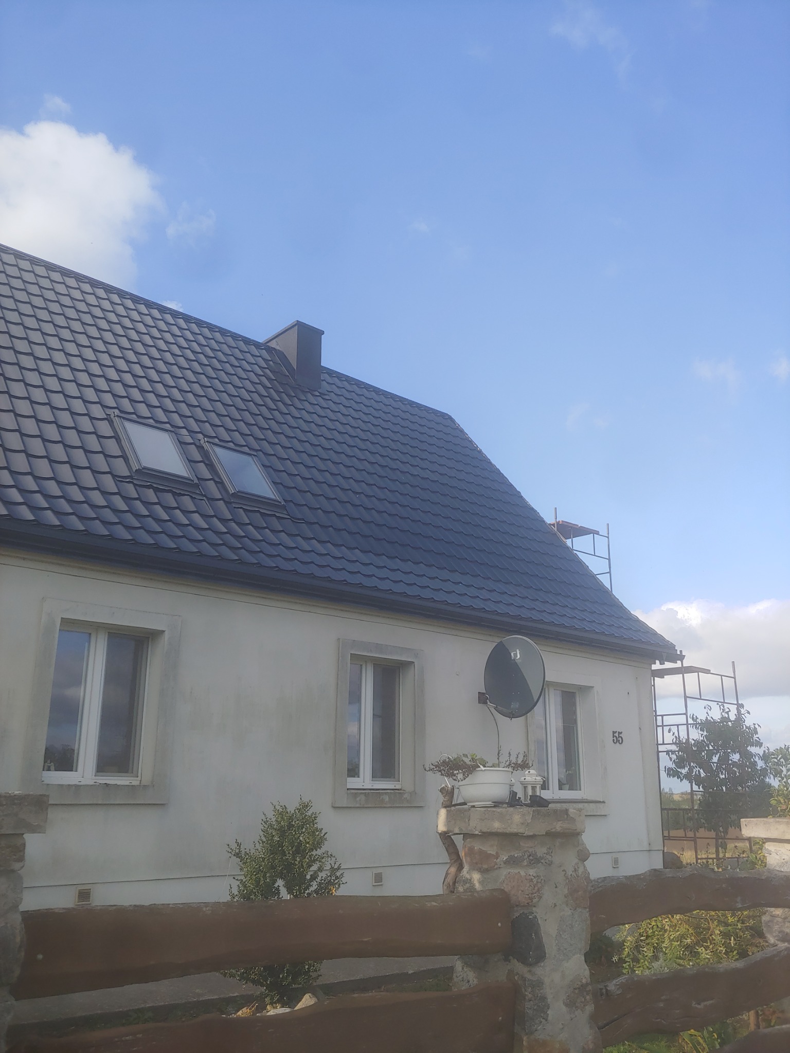 Dom jednorodzinny z nowym, ciemnym dachem z dachówki ceramicznej, widoczne dwa okna dachowe, antena satelitarna na ścianie i rusztowanie w tle.