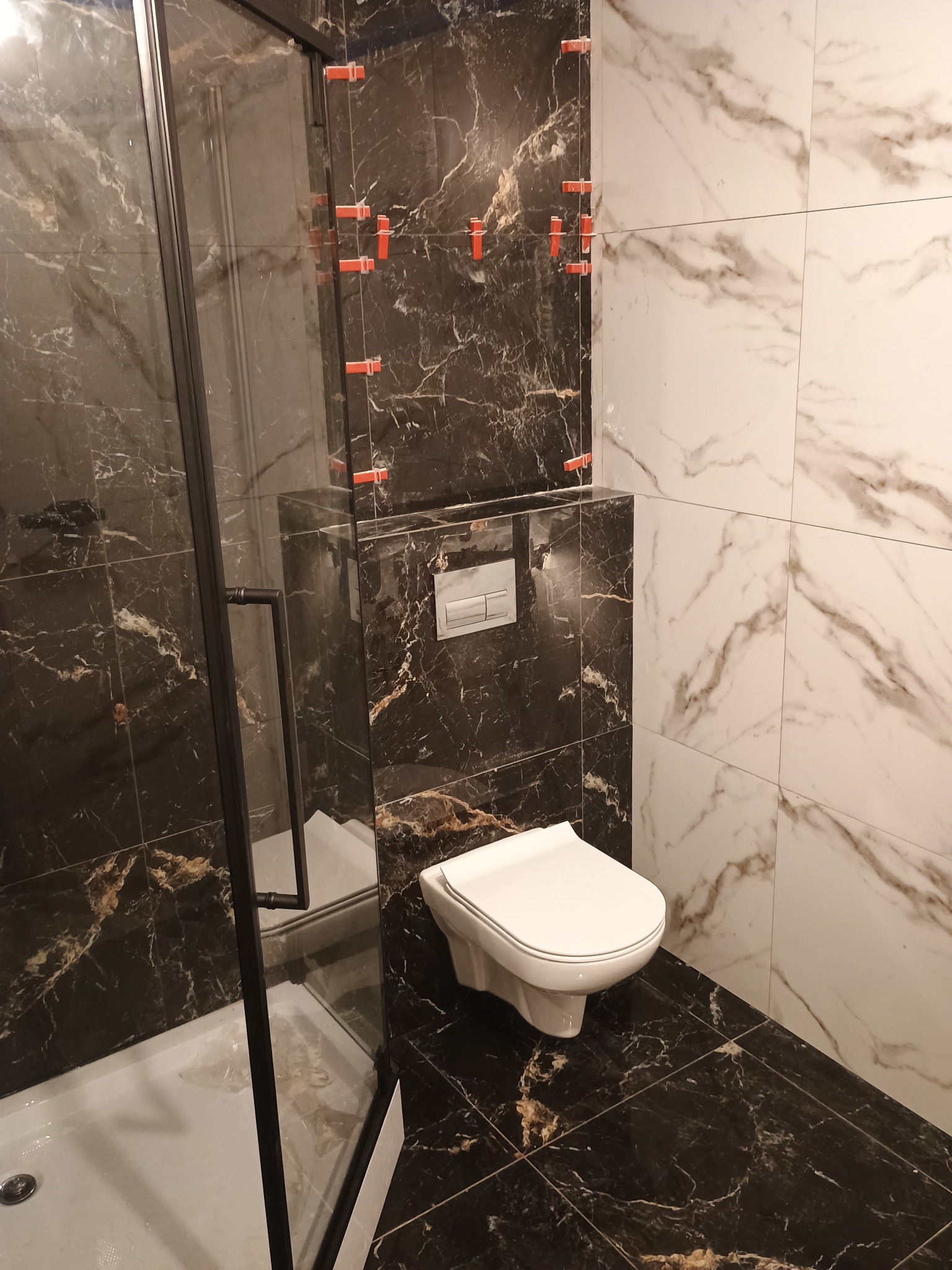 Nowoczesna łazienka w trakcie wykańczania: kabina prysznicowa z czarnymi profilami, wisząca toaleta i kombinacja płytek marmurowych w odcieniach bieli i czerni, z widocznymi klipsami poziomującymi...