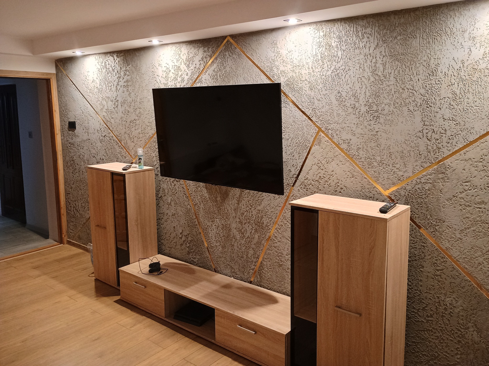 Szara ściana z dekoracyjną strukturą i złotymi geometrycznymi liniami, podświetlona punktowymi lampami sufitowymi, z zawieszonym telewizorem i meblami w odcieniach drewna.