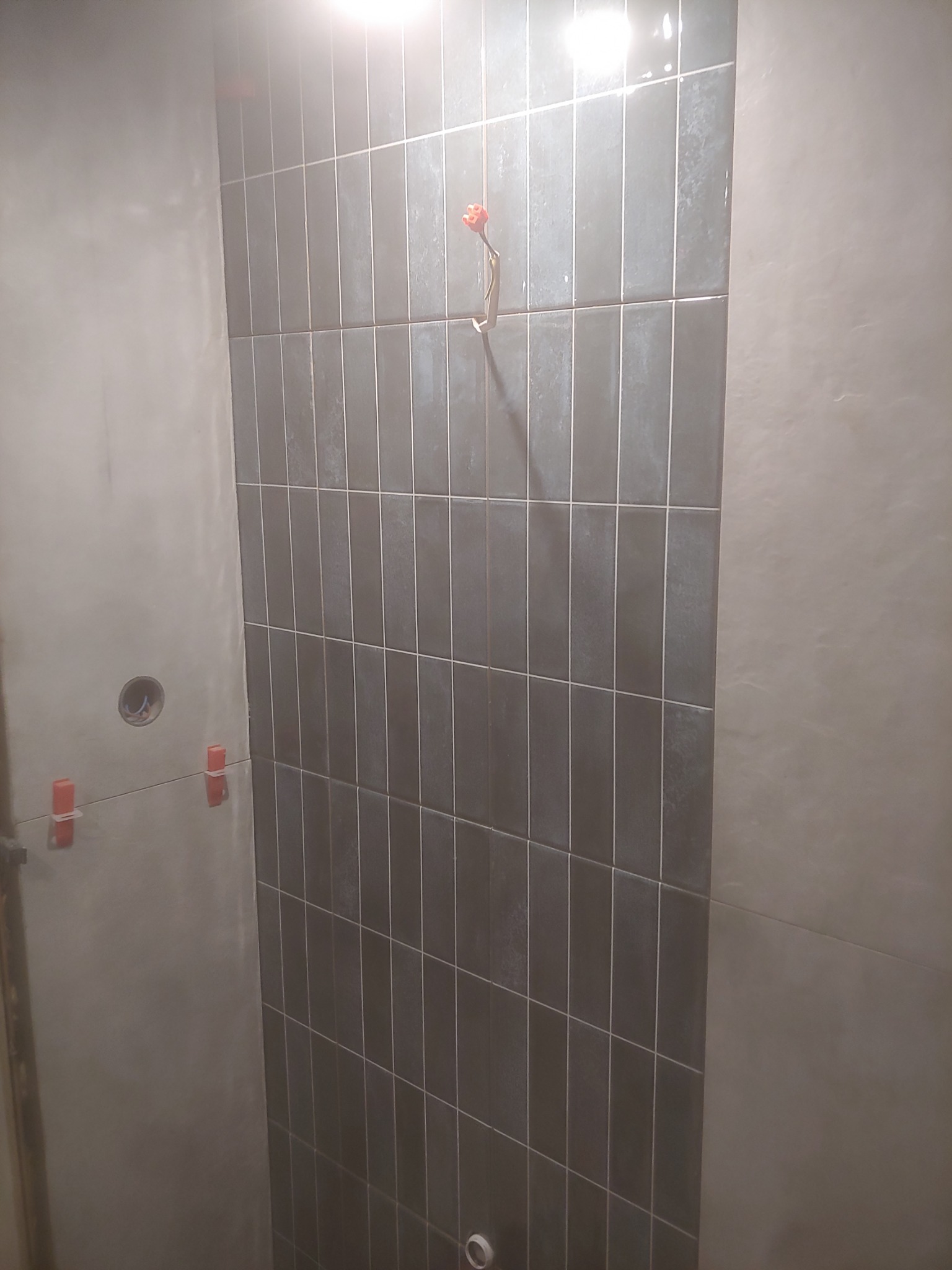 Układanie pionowych, grafitowych płytek w łazience, widoczne krzyżyki dystansowe, surowy tynk na ścianie obok.