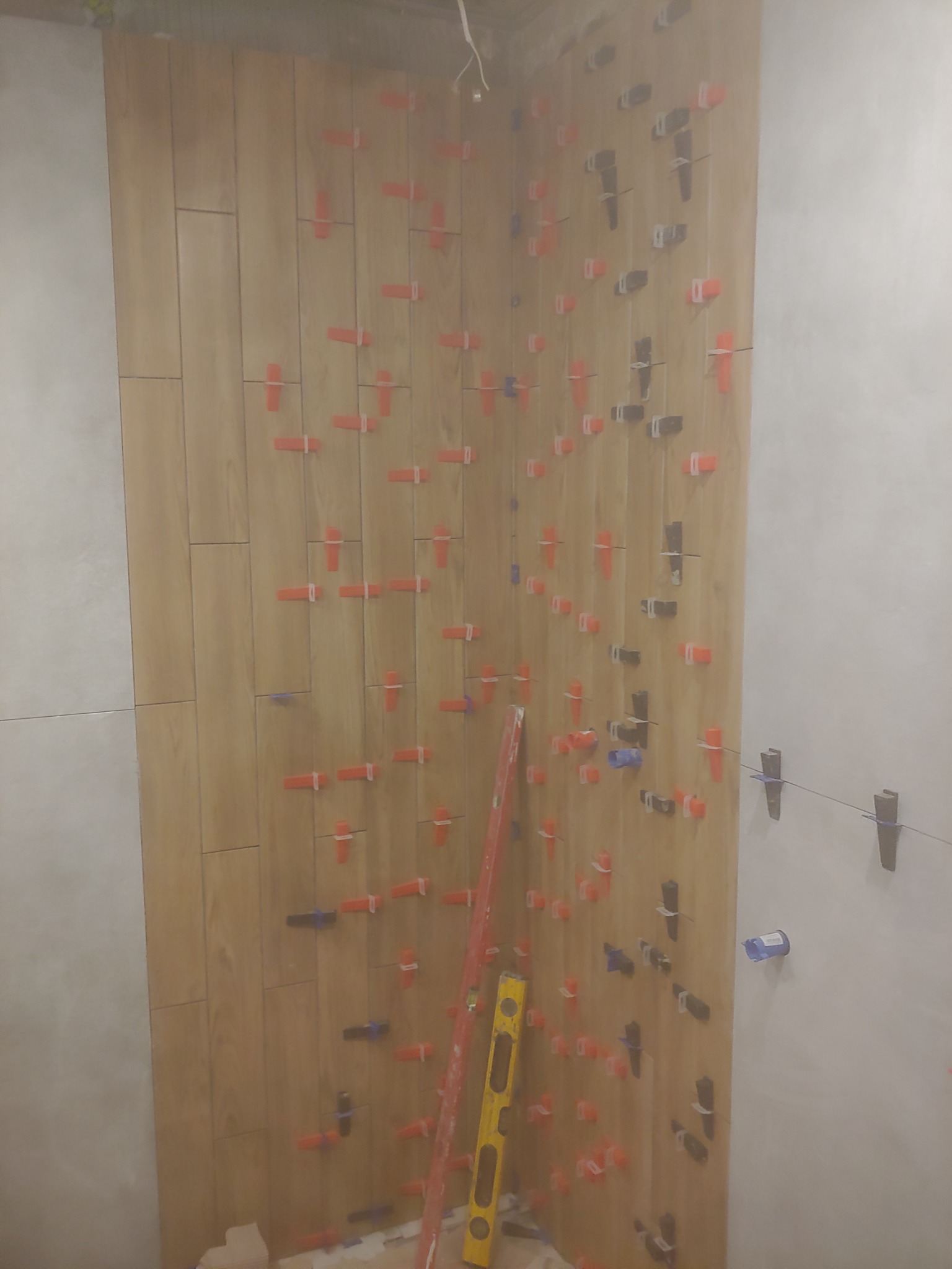 Ściana wyłożona drewnopodobnymi płytkami w trakcie montażu, z widocznymi krzyżykami dystansowymi w kolorach pomarańczowym, szarym i niebieskim, oraz pionową poziomicą przy krawędzi.