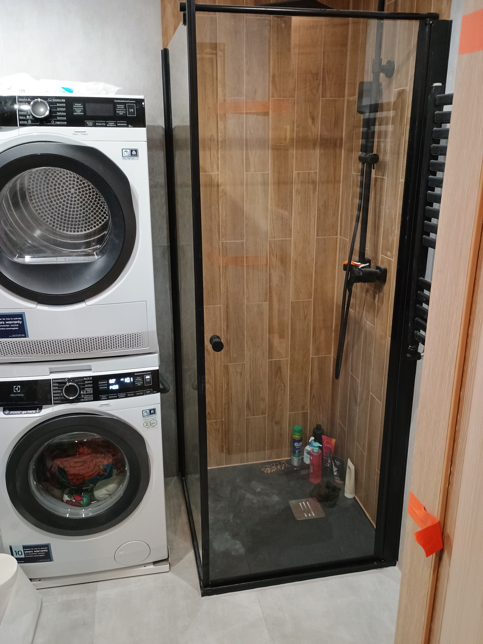 Nowoczesna łazienka z kabiną prysznicową typu walk-in z czarną armaturą i drewnopodobnymi płytkami, obok pralka i suszarka Electrolux ustawione jedna na drugiej.