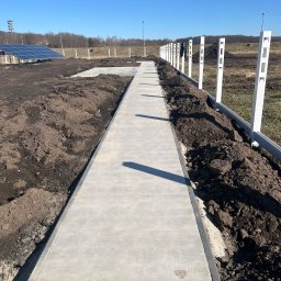Pep-Bud - Długa, prosta betonowa ścieżka wzdłuż białego ogrodzenia panelowego na nierównym, przygotowywanym terenie, panele słoneczne widoczne w tle.