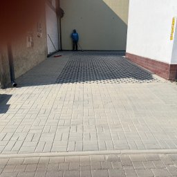 Pep-Bud - Ułożona jasnoszara kostka brukowa na podjeździe, z fragmentem kostki ażurowej w tle. Pracownik zamiata. Widoczne budynki po bokach.