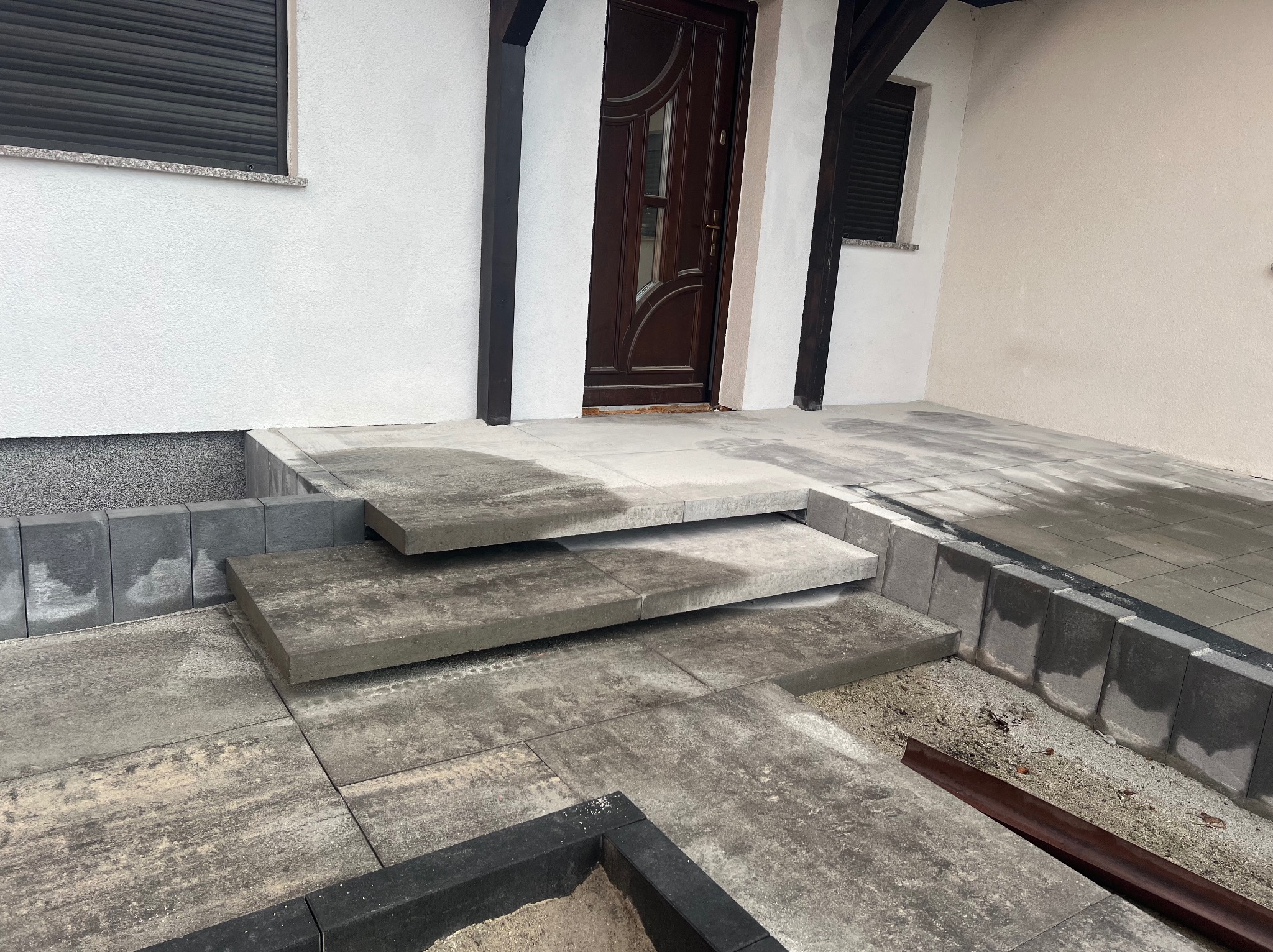 Ułożona kostka brukowa tworząca schody wejściowe do domu z brązowymi drzwiami i białymi ścianami. Widoczne obrzeża i elementy wykończeniowe.