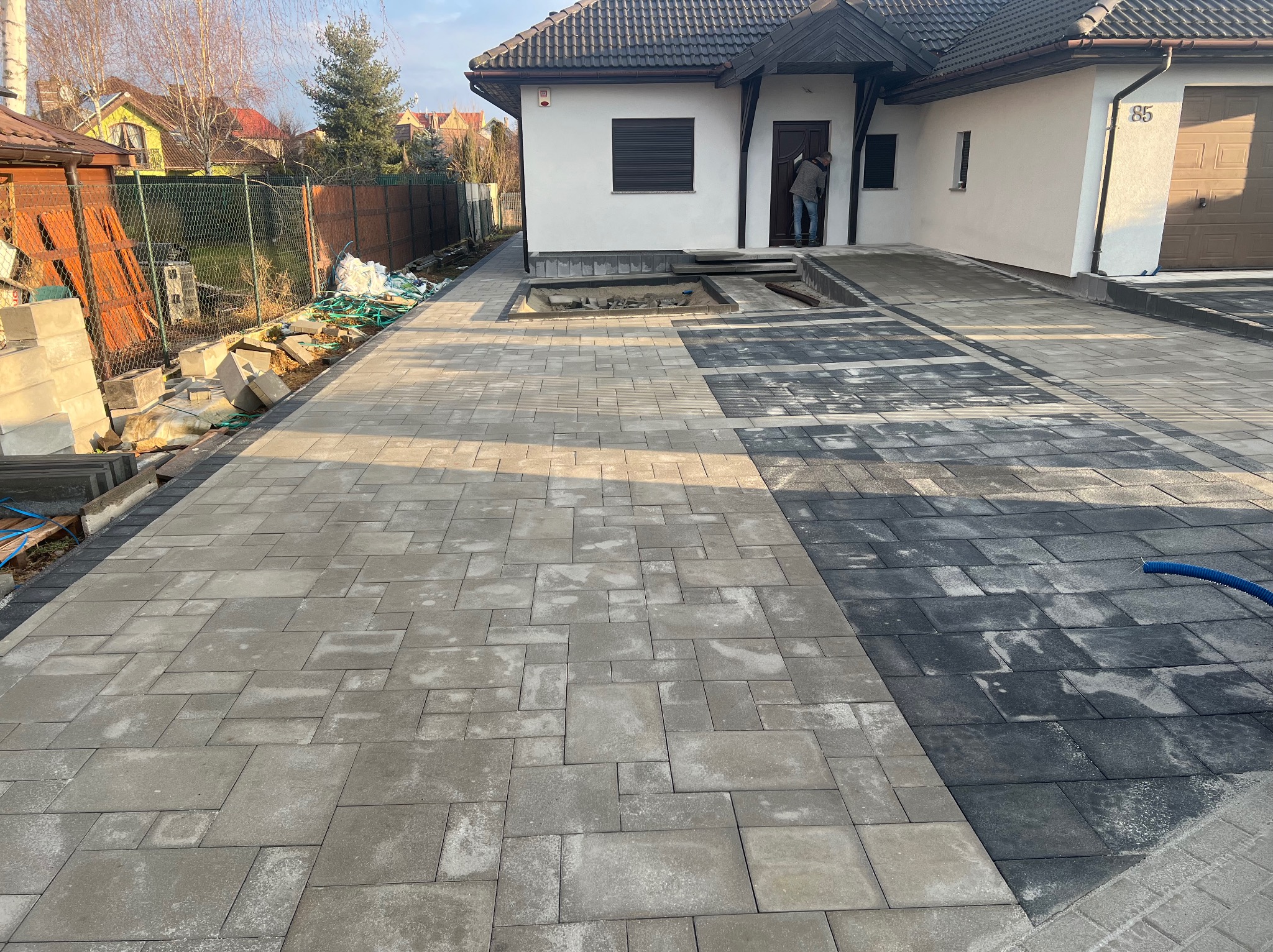 Ułożona kostka brukowa w dwóch odcieniach szarości prowadząca do domu z numerem 85, z widocznym fragmentem przygotowywanego podłoża pod rabatę kwiatową i osobą wchodzącą do domu.
