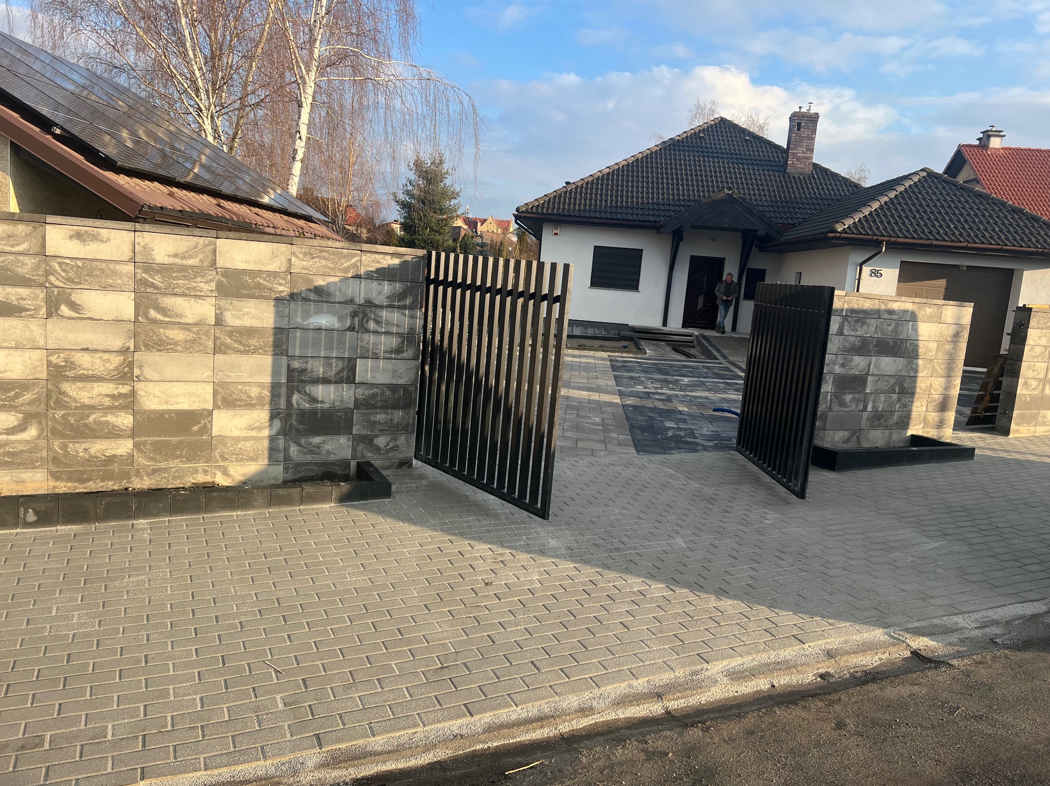 Nowoczesna posesja z szarej kostki brukowej prowadząca do domu z otwartą, metalową bramą i murem z betonowych bloczków.