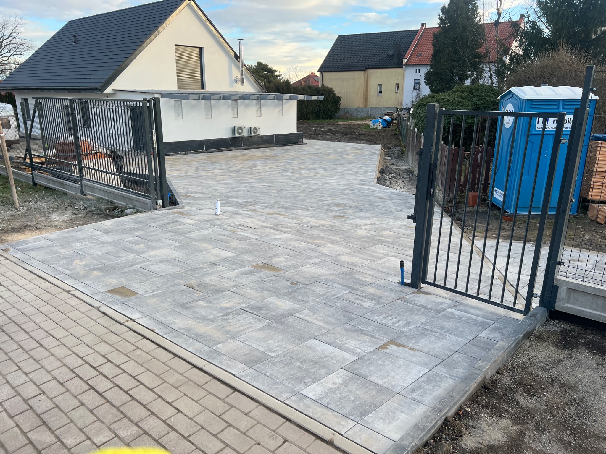 Ułożona szara kostka brukowa na podjeździe, widoczne otwarte metalowe bramy wjazdowe, w tle dom jednorodzinny i przenośna toaleta.