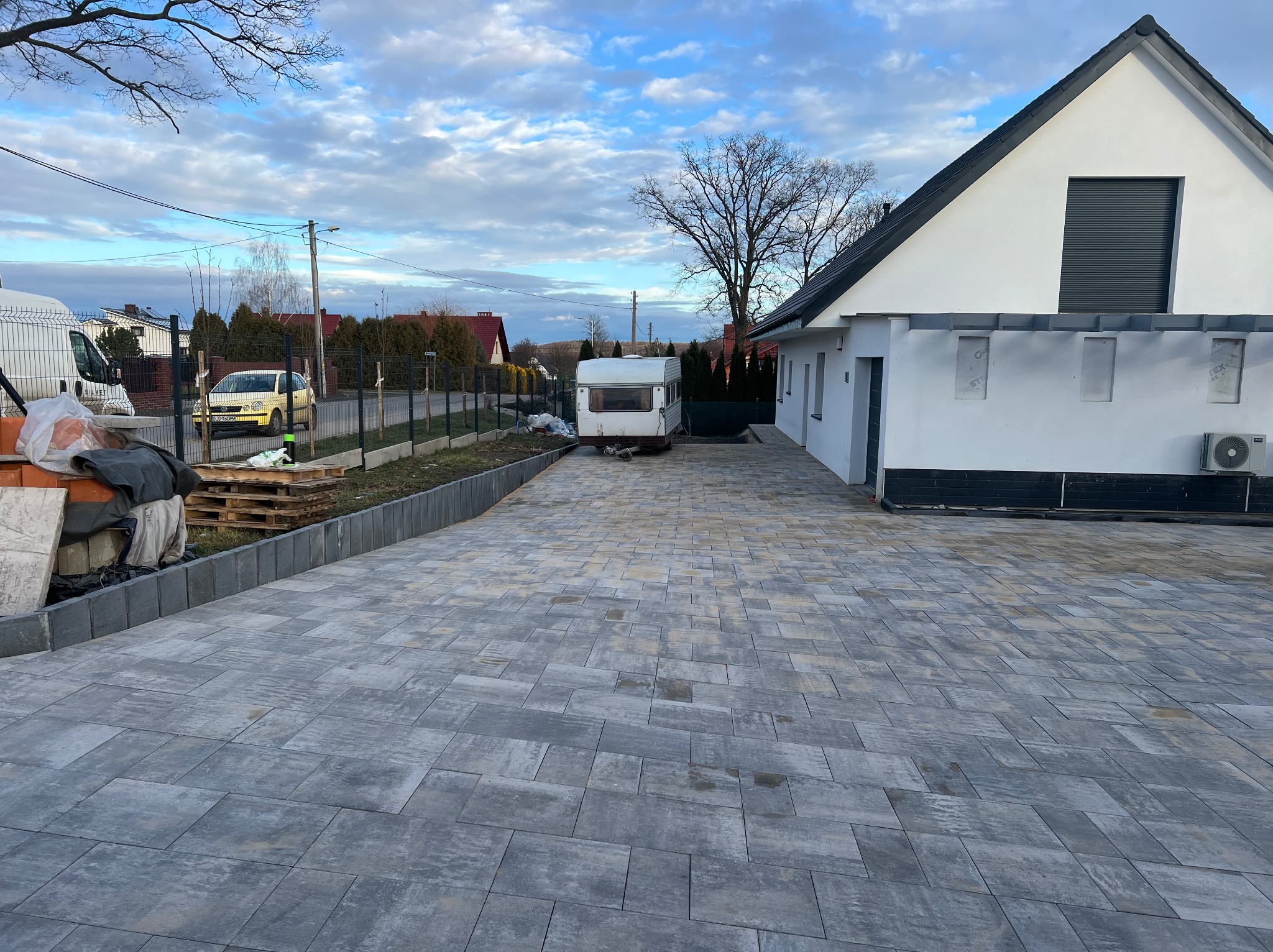 Szara kostka brukowa ułożona na podjeździe do domu z białymi ścianami i szarym dachem, z zaparkowaną przyczepą kempingową w tle i widocznymi paletami z materiałami budowlanymi po lewej stronie.