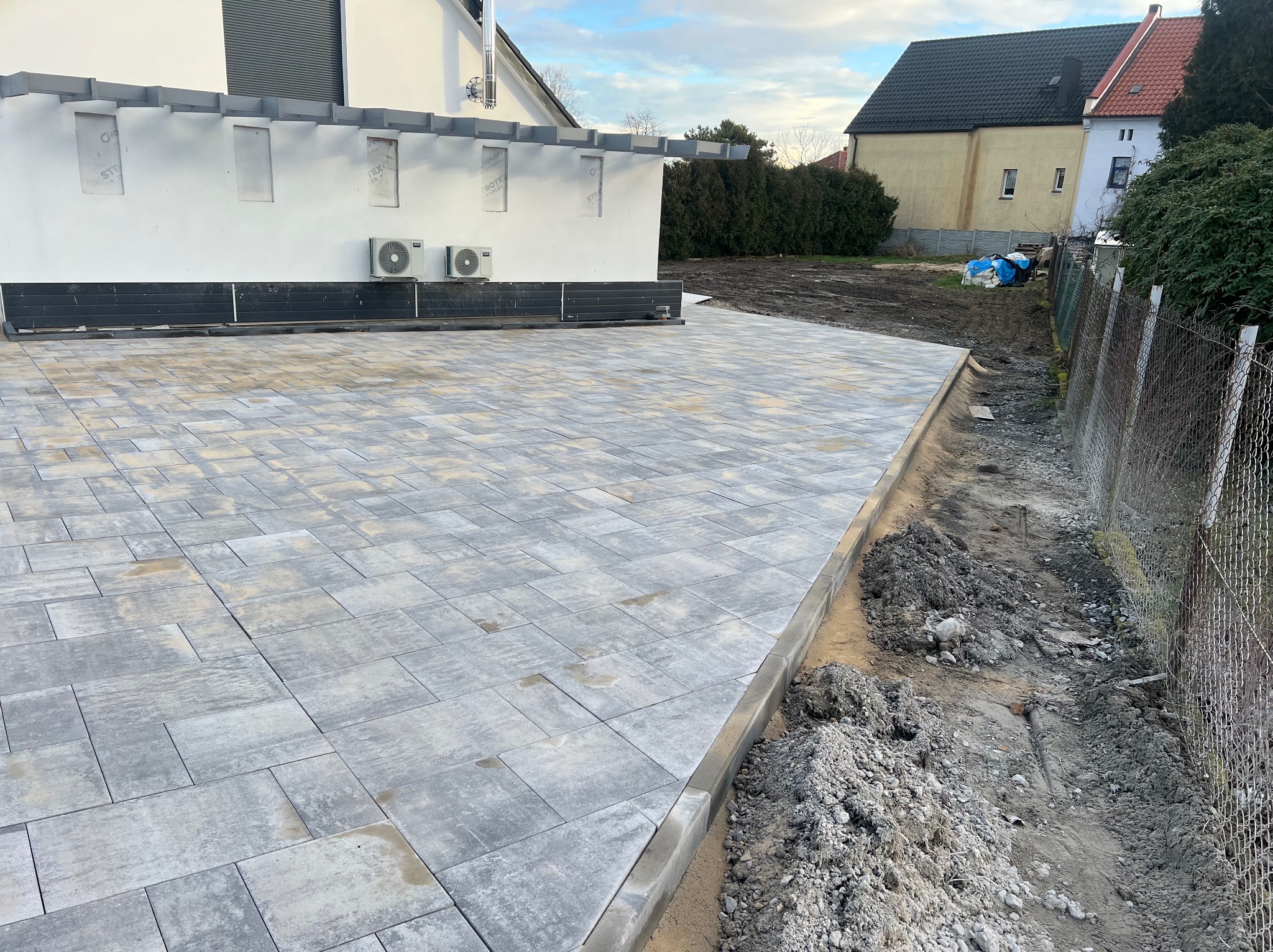 Nowa, szara nawierzchnia z kostki brukowej ułożona przed nowoczesnym domem z białymi ścianami i klimatyzatorami, widoczna granica z ziemią i ogrodzeniem z siatki.