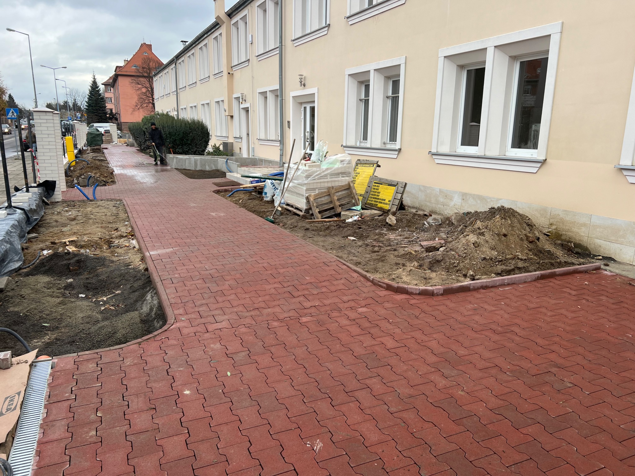 Czerwona kostka brukowa ułożona wzdłuż budynku z jasną elewacją, widoczne przygotowania do dalszych prac brukarskich, palety z materiałami budowlanymi i narzędzia.