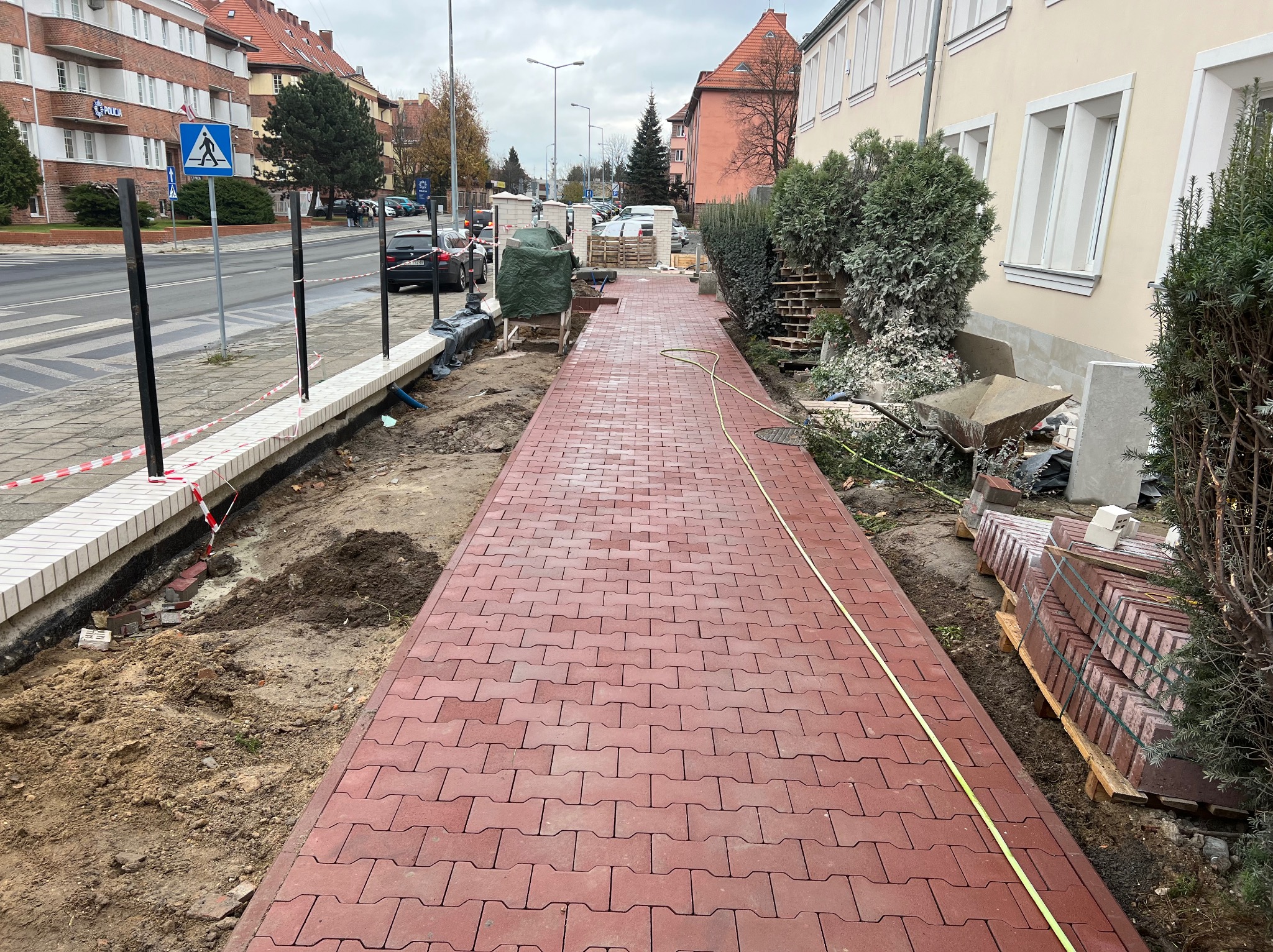 Czerwona, ułożona ścieżka z kostki brukowej wzdłuż ulicy miejskiej, w tle budynki mieszkalne i znak drogowy dla pieszych. Po lewej stronie widoczny przygotowany podkład pod dalszą część chodnika...