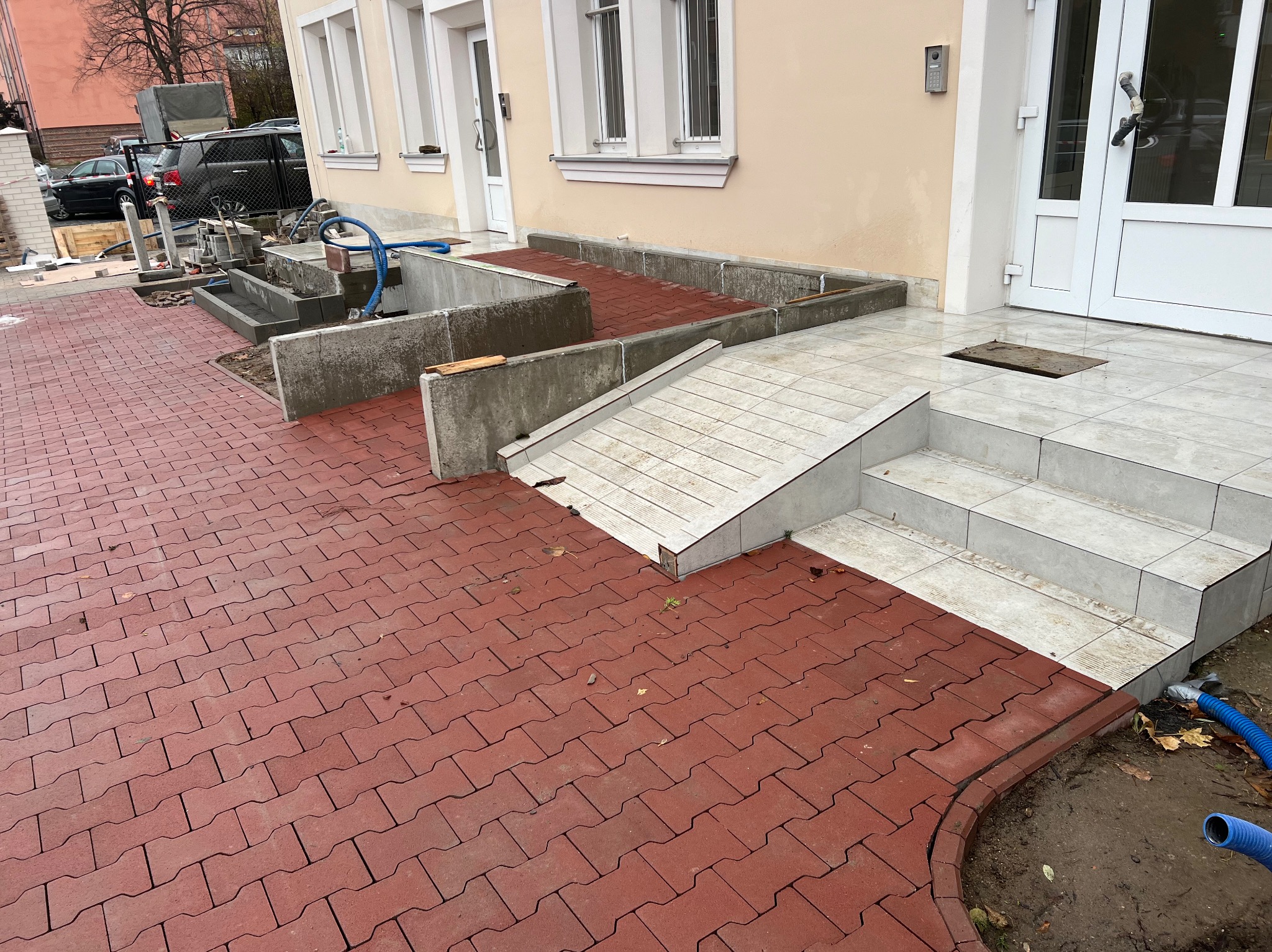 Czerwona kostka brukowa ułożona przed budynkiem z rampą i schodami, widoczne elementy betonowe w trakcie prac wykończeniowych.