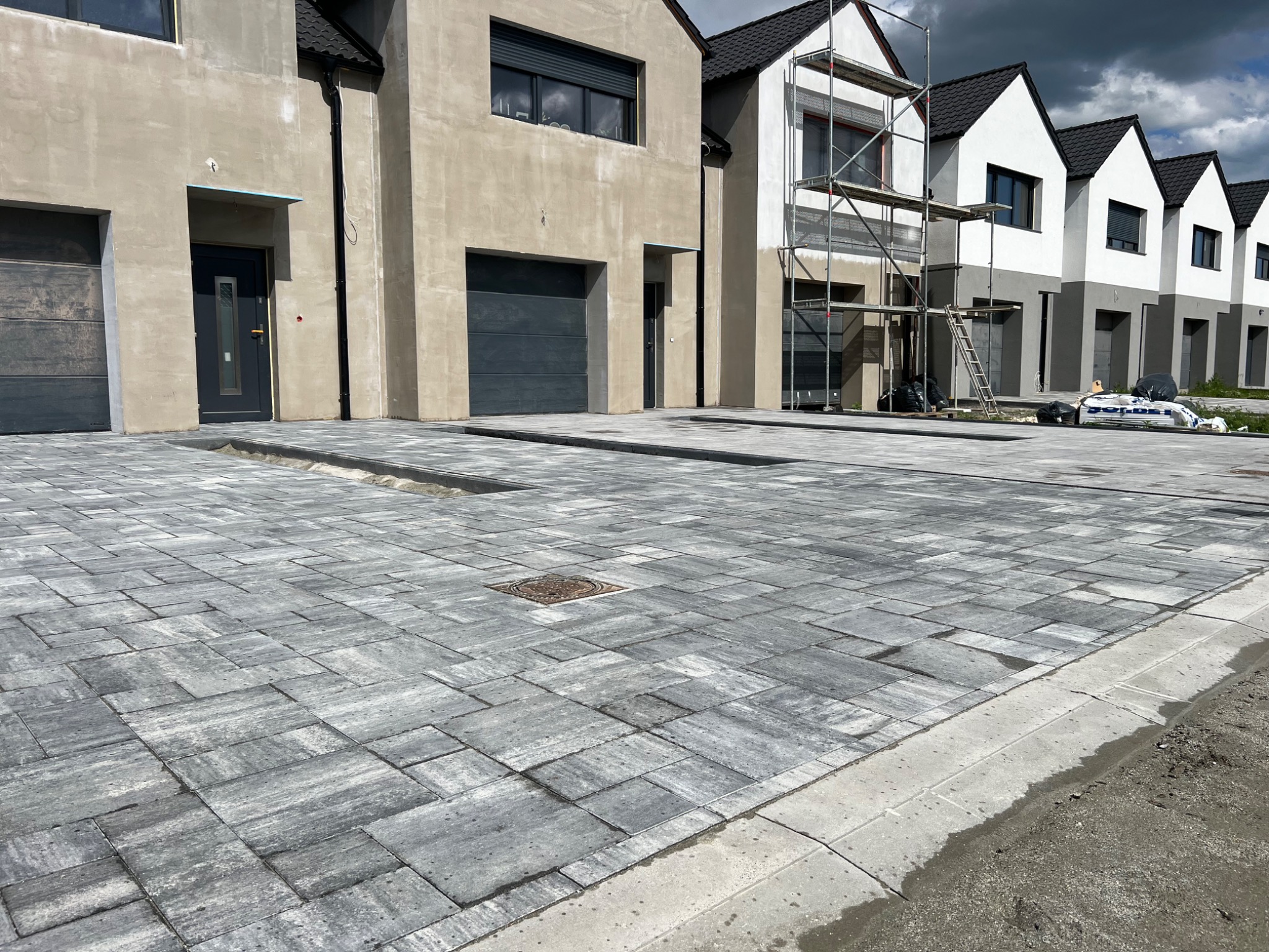 Szara kostka brukowa ułożona na podjeździe do szeregu nowych domów, z widoczną studzienką kanalizacyjną. Część domów w trakcie wykończenia elewacji.