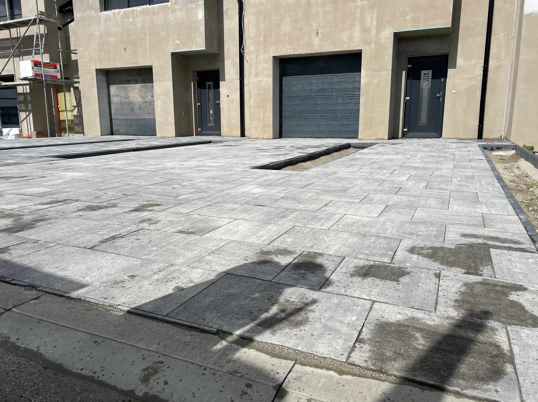 Nowa, szara kostka brukowa ułożona przed budynkiem z trzema garażami. Widoczne odpływy liniowe. Częściowo mokra nawierzchnia z odbiciem cienia.
