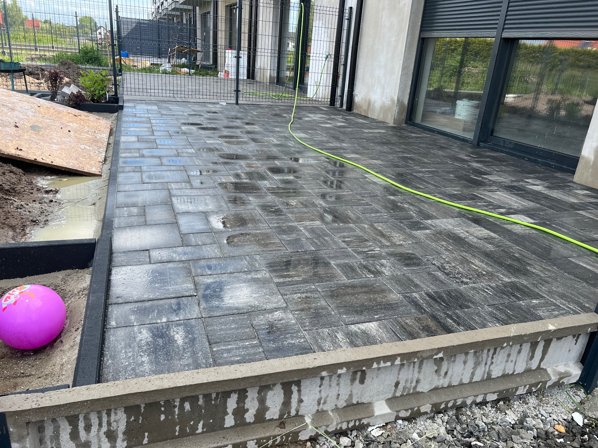 Ułożona, mokra kostka brukowa w szarych odcieniach na posesji z metalowym ogrodzeniem. W tle widoczny budynek mieszkalny, a obok kostki leży różowa piłka.
