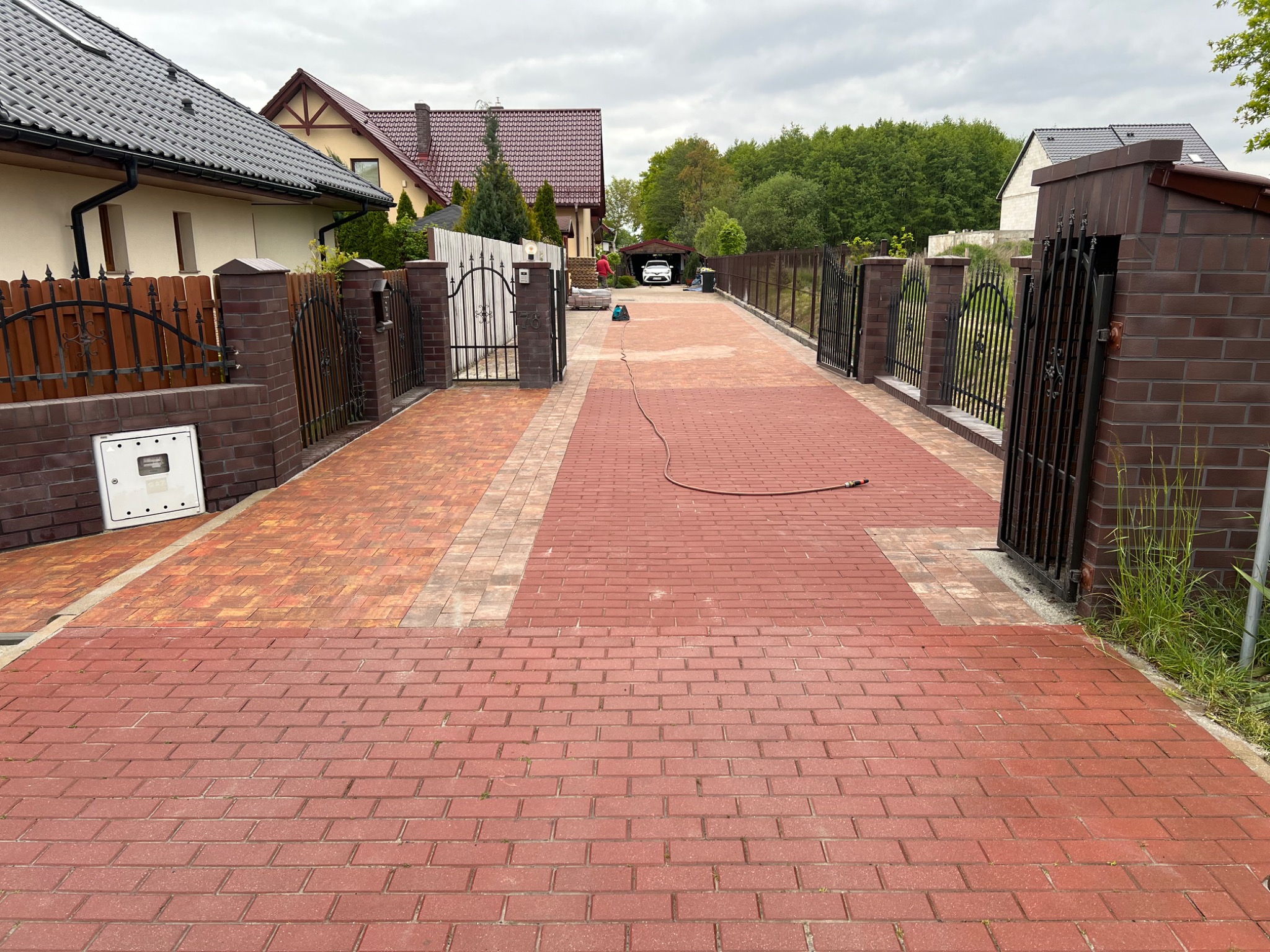 Czerwona kostka brukowa na podjeździe, brama z kutego żelaza i ceglane słupki ogrodzeniowe, domy w tle, kabel na kostce brukowej.