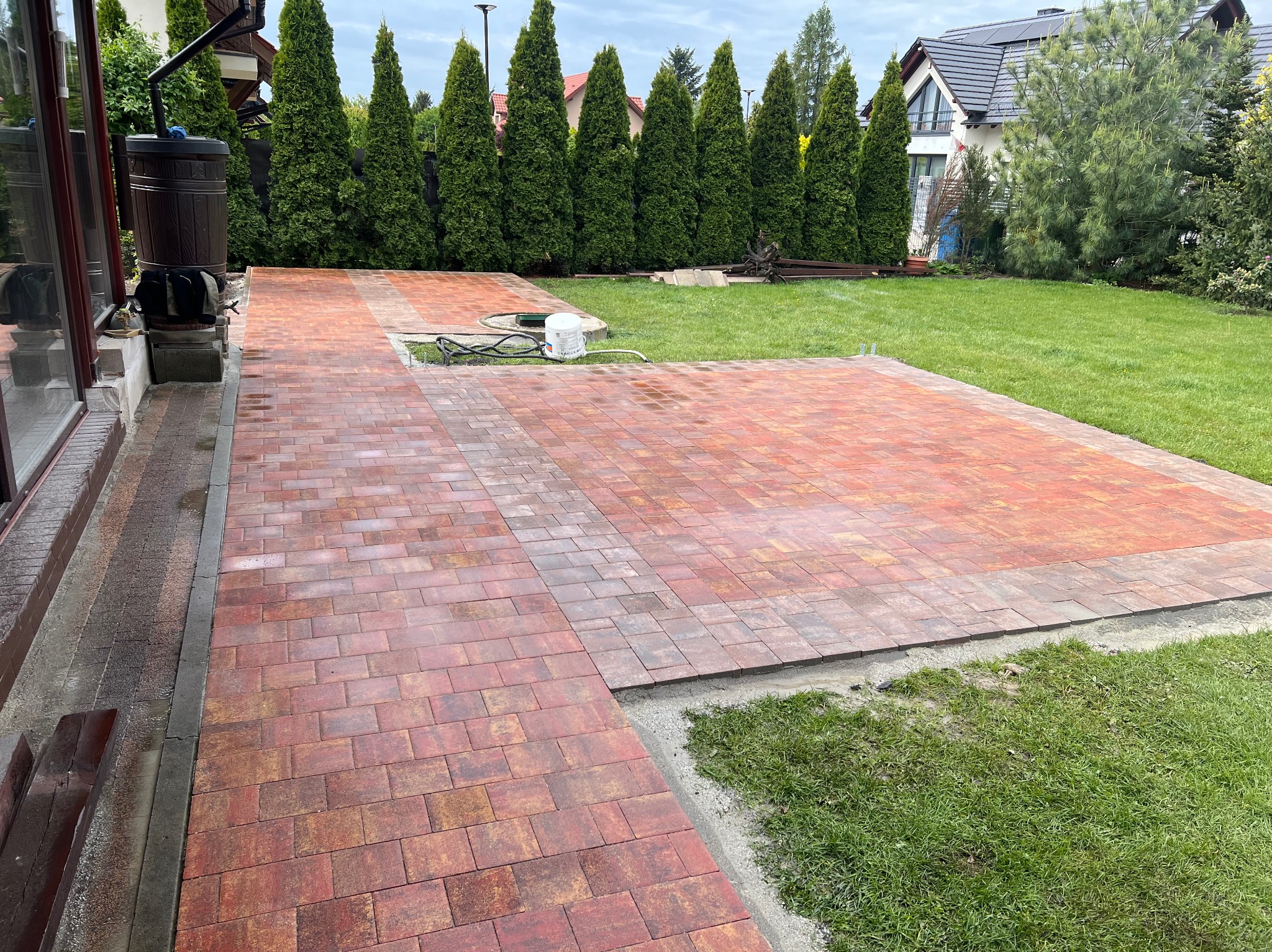 Nowa, czerwono-brązowa kostka brukowa na posesji, częściowo ułożona, z widocznymi elementami przygotowania podłoża i zielonym trawnikiem w tle.