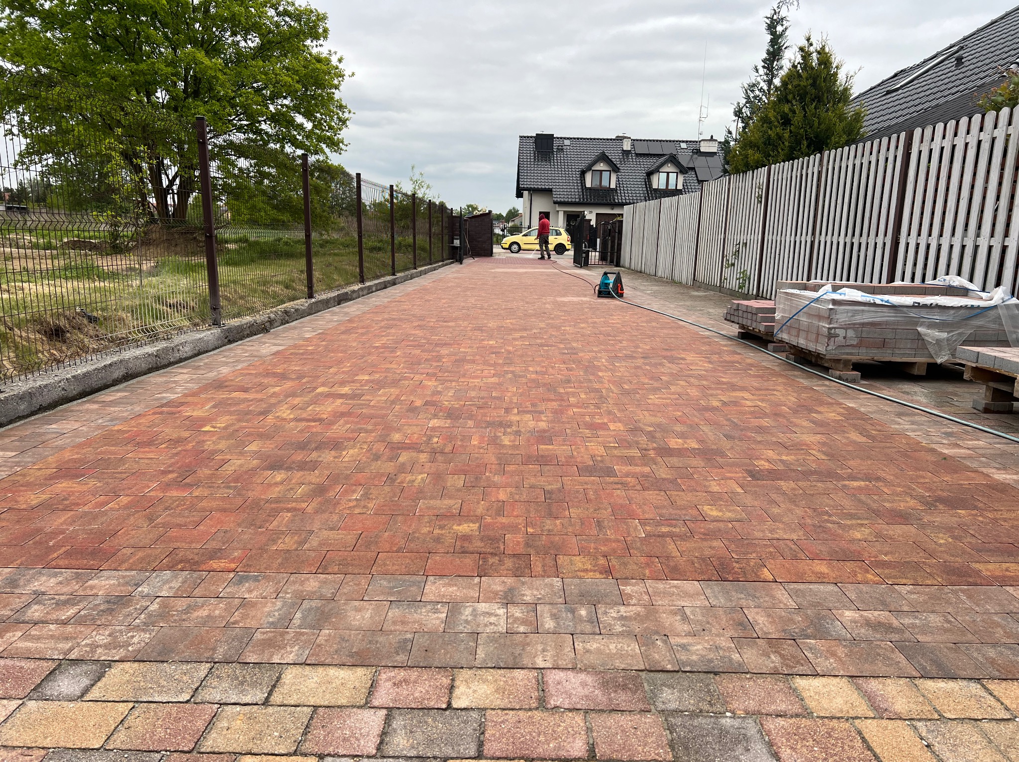 Ukończony podjazd z kostki brukowej w odcieniach czerwieni i brązu, otoczony ogrodzeniem panelowym i drewnianym płotem, z widocznym samochodem w tle.