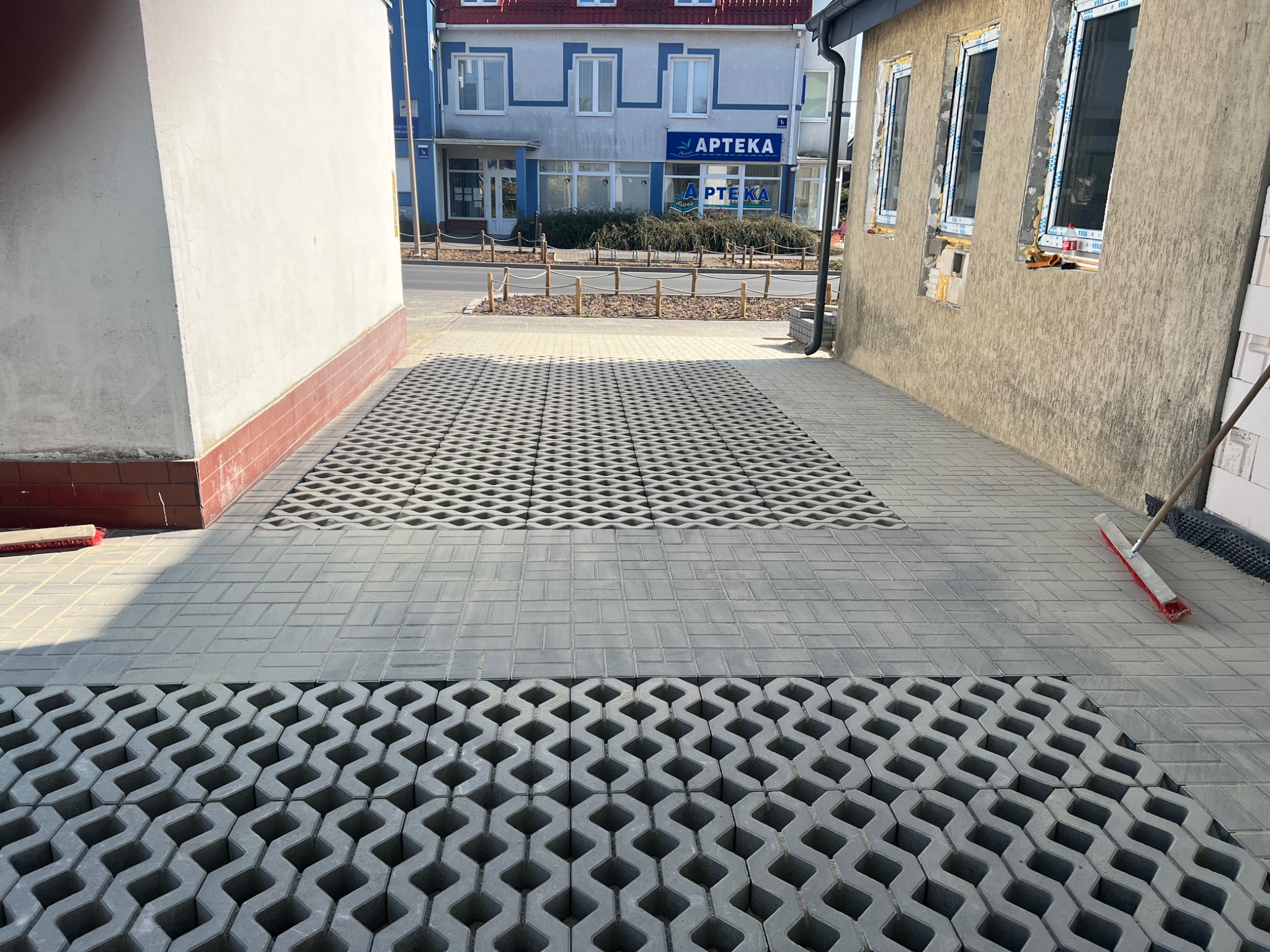 Szara, częściowo ułożona kostka ażurowa i brukowa na podjeździe między budynkami. W tle widoczny budynek apteki i fragment ściany z oknami.