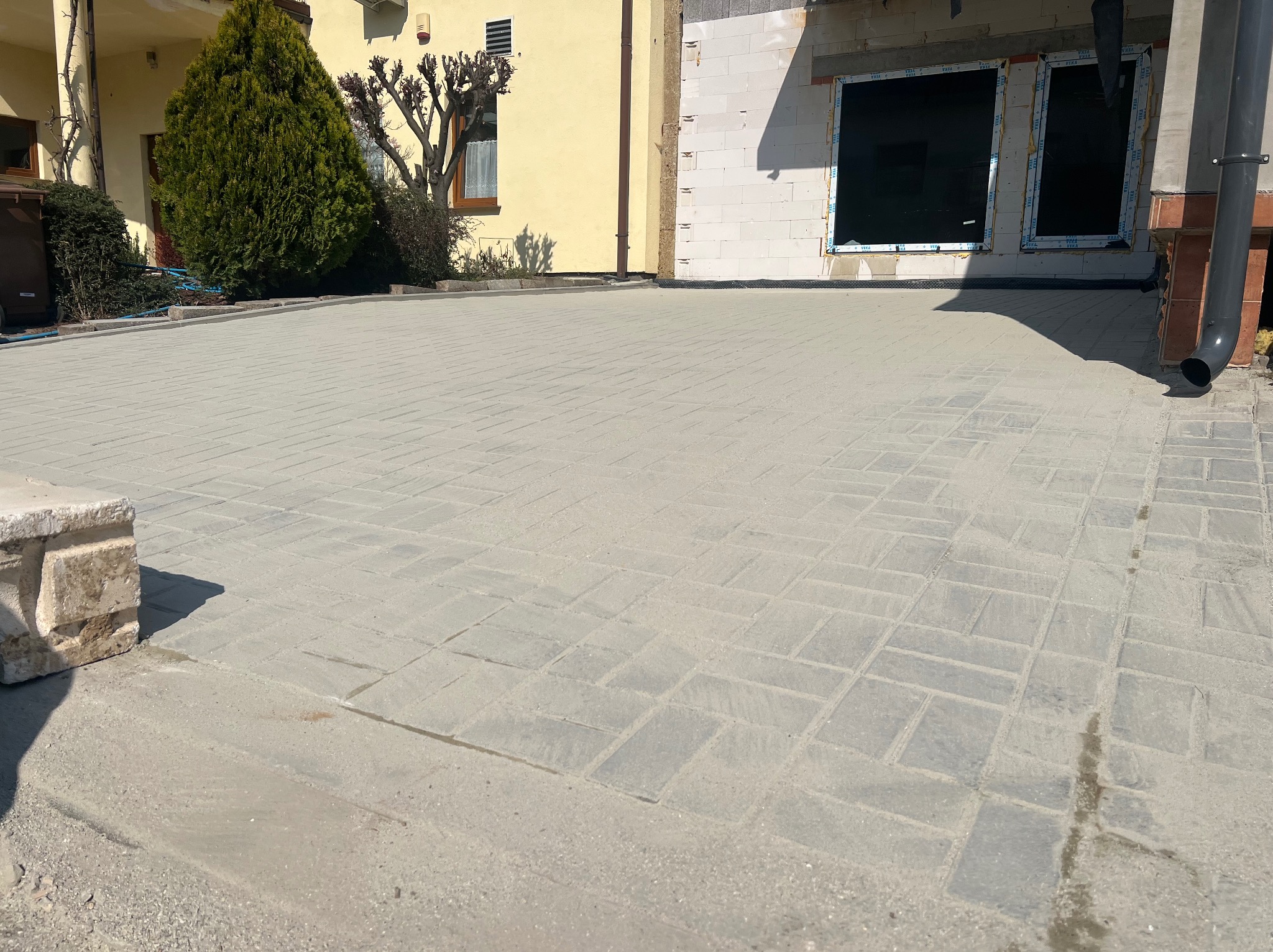 Jasnoszara kostka brukowa ułożona na podjeździe przed domem w słoneczny dzień. Widoczny fragment budynku w trakcie budowy i elementy zieleni.