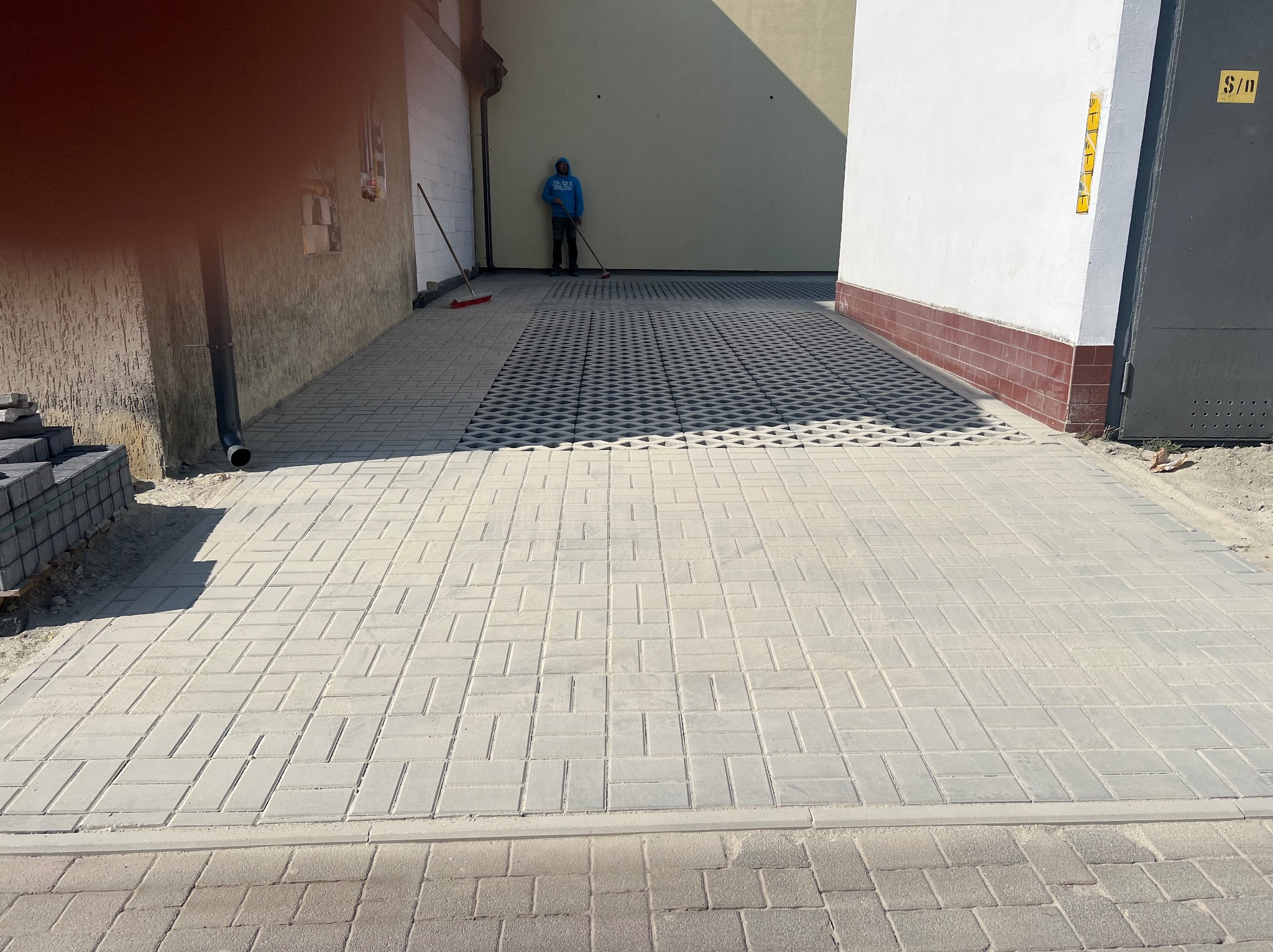 Ułożona jasnoszara kostka brukowa na podjeździe, z fragmentem kostki ażurowej w tle. Pracownik zamiata. Widoczne budynki po bokach.