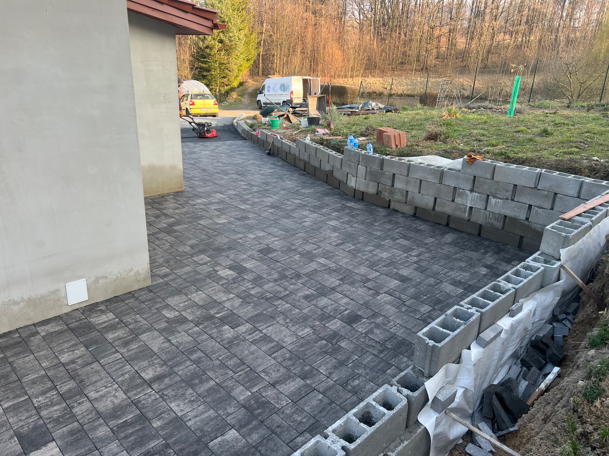 Częściowo ułożona kostka brukowa w odcieniach szarości, obramowana murem z bloczków betonowych. W tle trawnik, drzewa i budynek. Widoczny zagęszczarka wibracyjna.